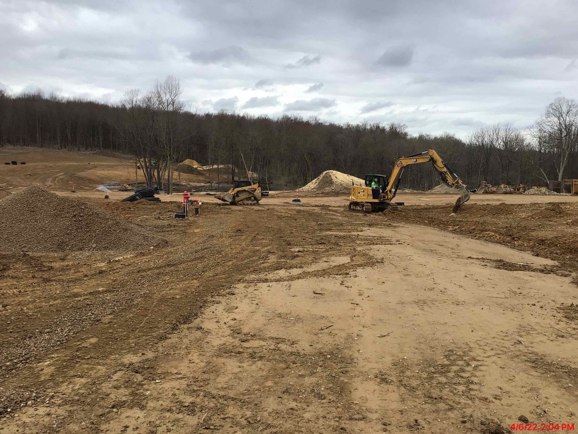 Construction site with excavators on muddy ground. Trees in the background. Overcast sky.