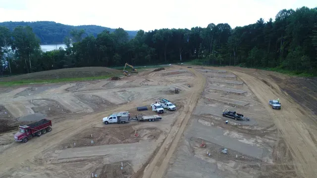Construction site with dirt roads, heavy machinery, and several trucks, with trees and water in background.