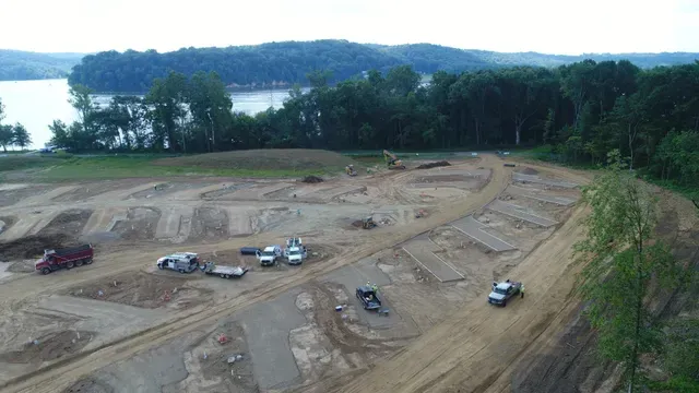 Construction site with dirt piles and vehicles near a body of water and treeline under cloudy skies.