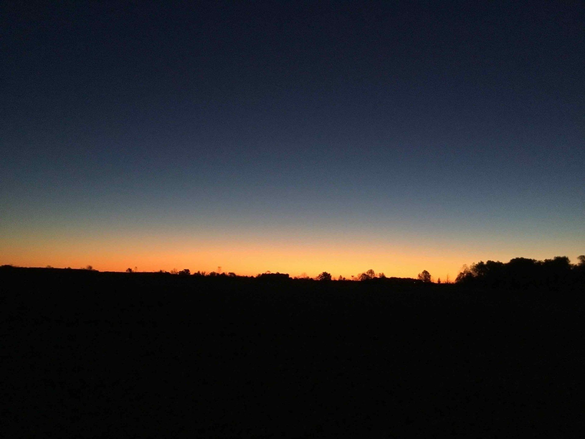 Sunset over a dark landscape, with orange and yellow hues at the horizon transitioning to a dark blue sky.