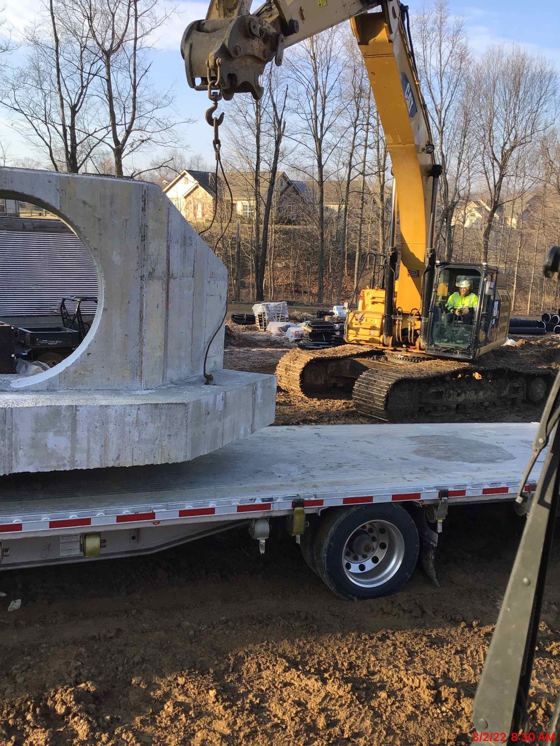 Excavator lifting a large concrete structure from a flatbed trailer at a construction site.