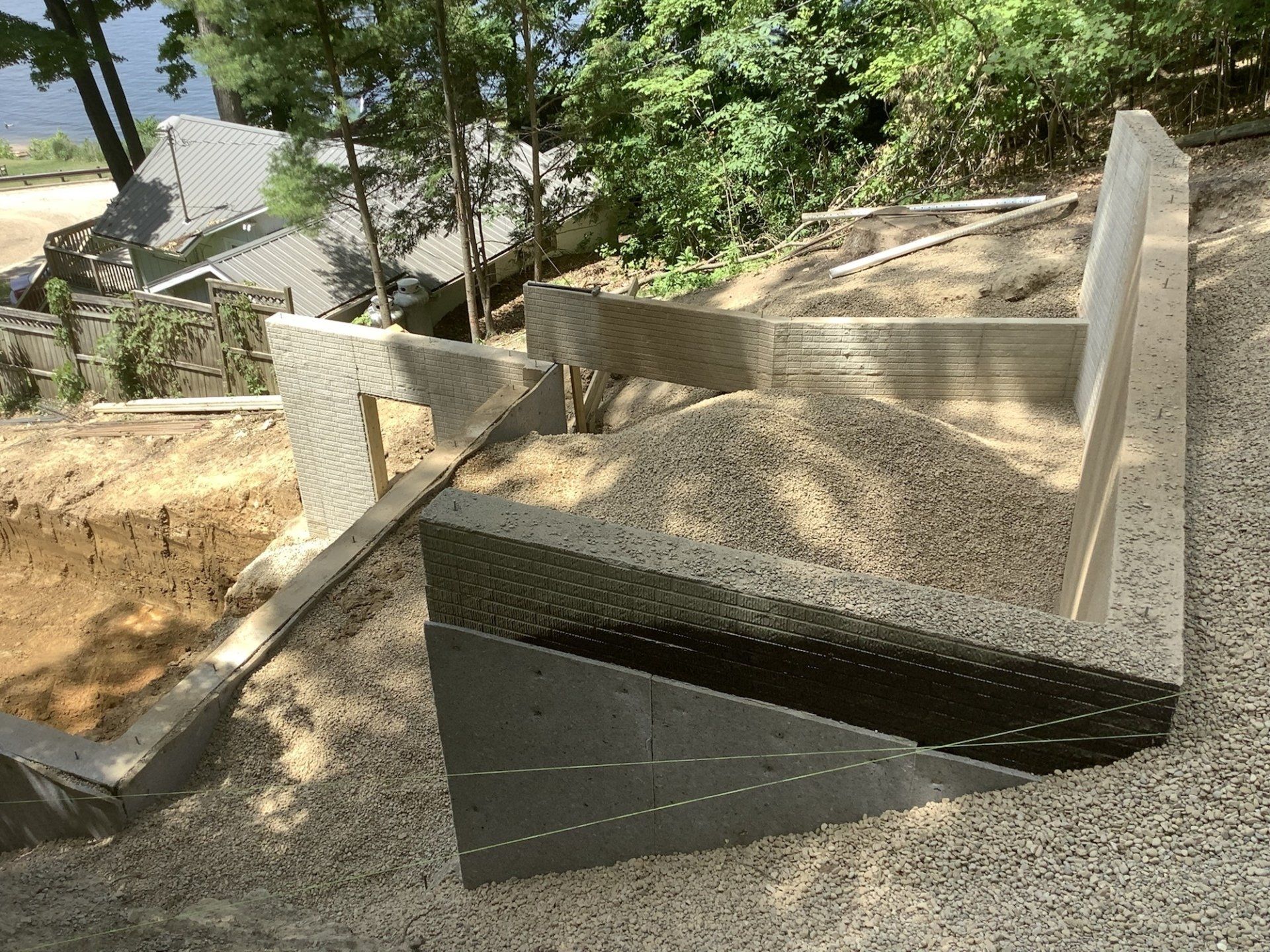 Retaining walls constructed on a hillside, filled with gravel. Green foliage and water visible in the background.