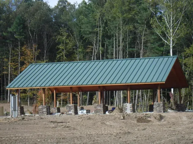 Green roofed pavilion under construction. Wooden posts and roof structure, surrounded by forest.