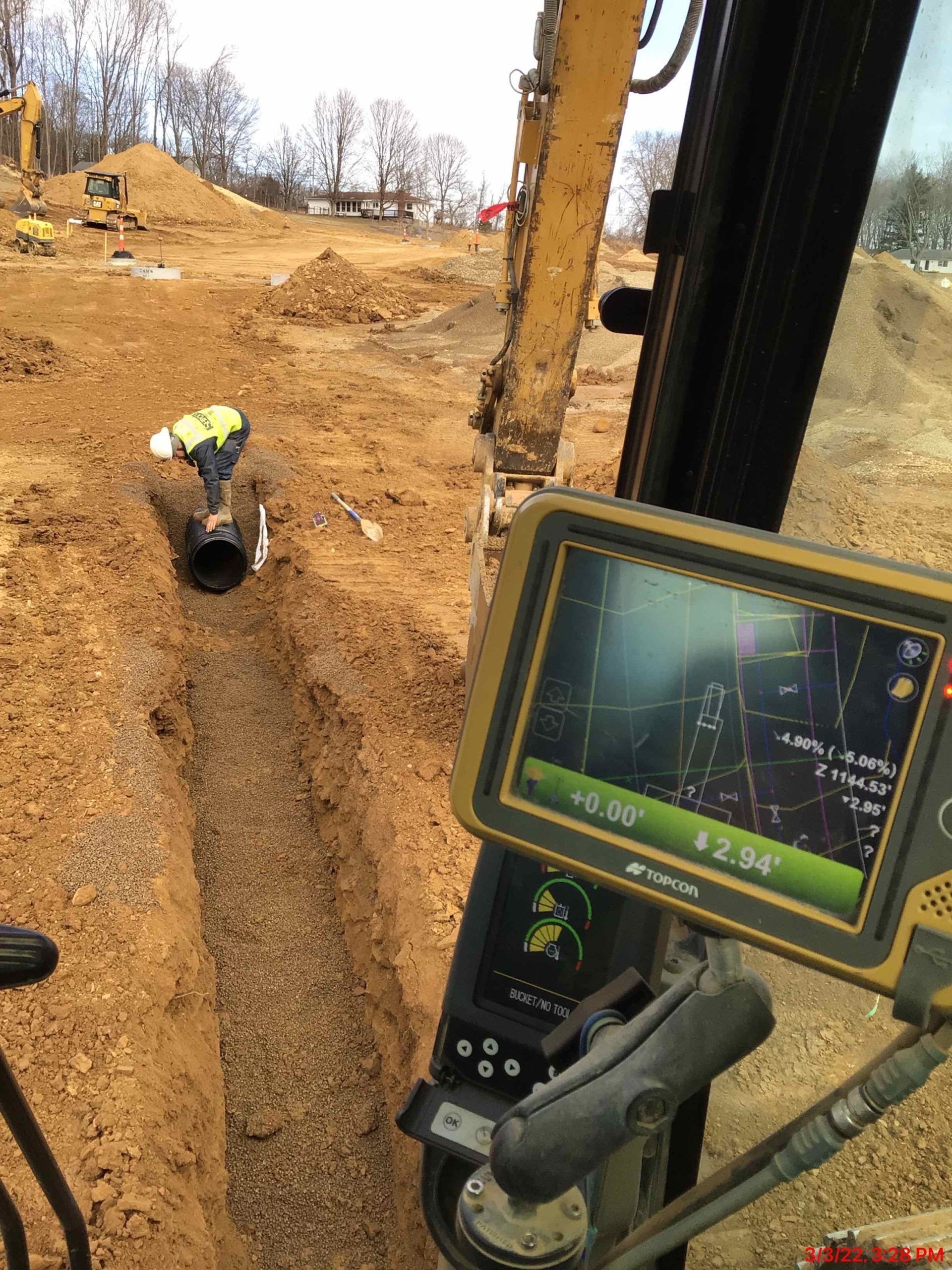 Excavator cab view; screen displays site map. Person works near pipe in trench. Construction site, daylight.