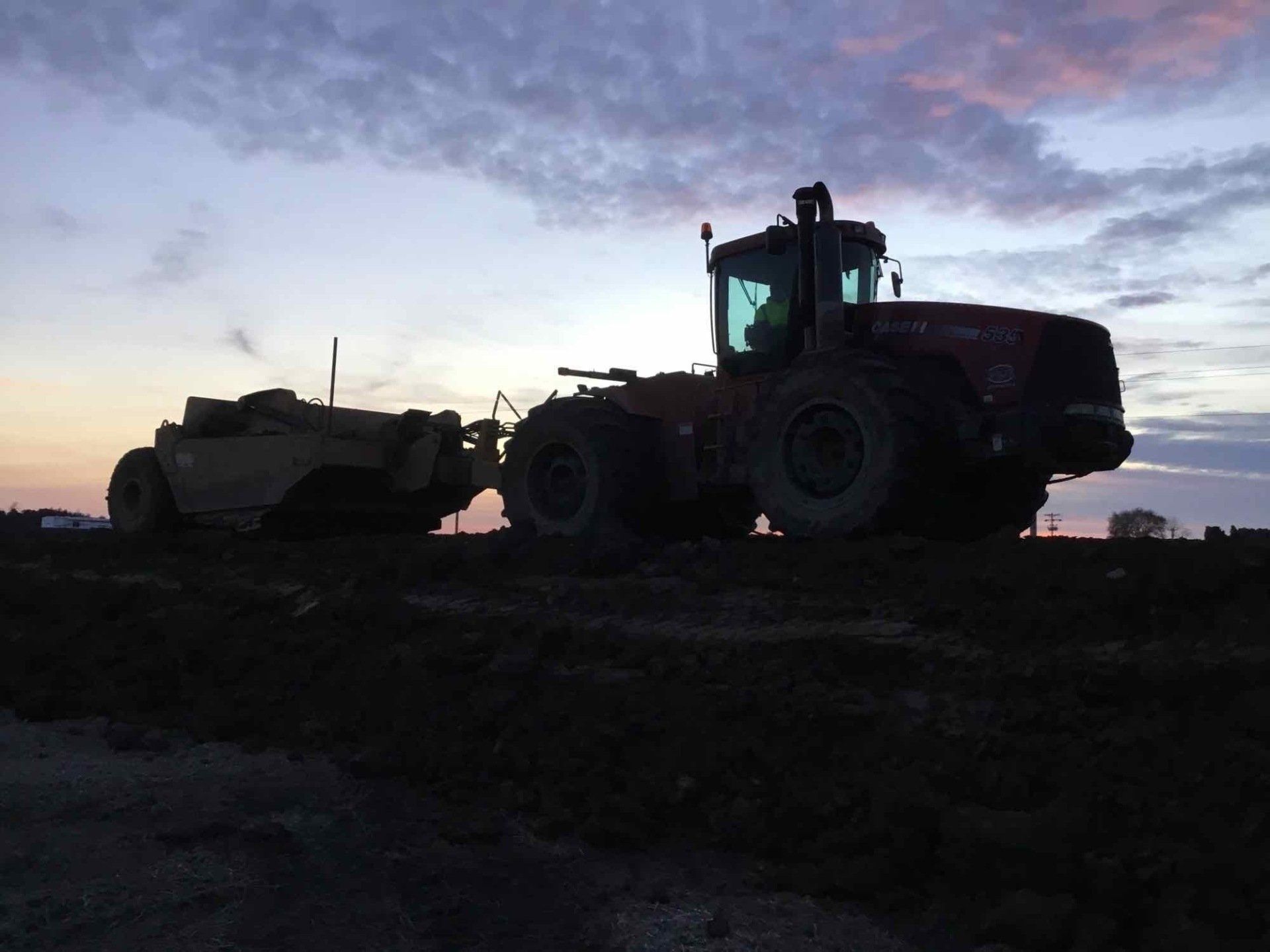 Large tractor pulling a piece of farm equipment across a dark field at dusk.