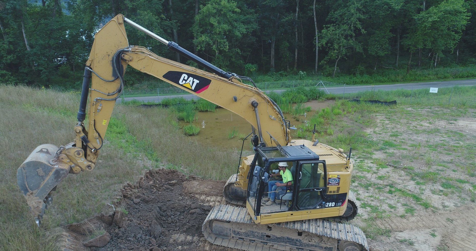 Yellow Caterpillar excavator digging earth. Operator visible in cab. Green trees in background.