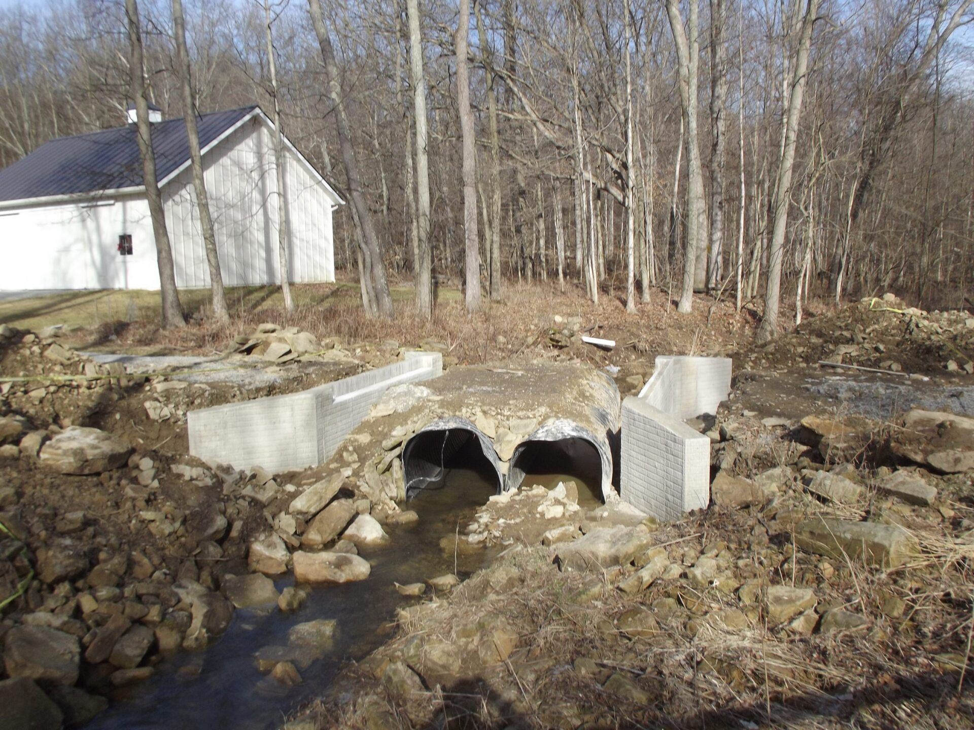 Damaged concrete culvert over a stream in a rural setting, near a white building.