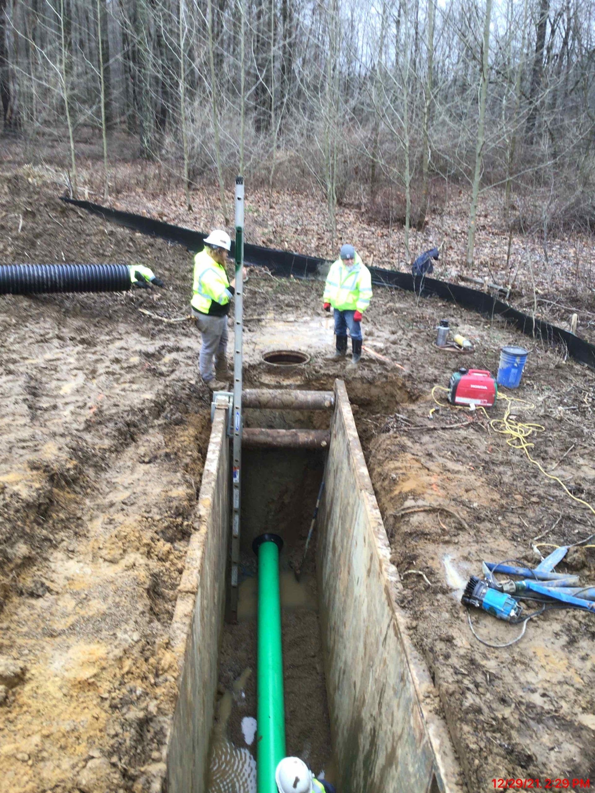Workers installing green pipe in a trench. One holds a level, another stands nearby. Outdoors with forest background.
