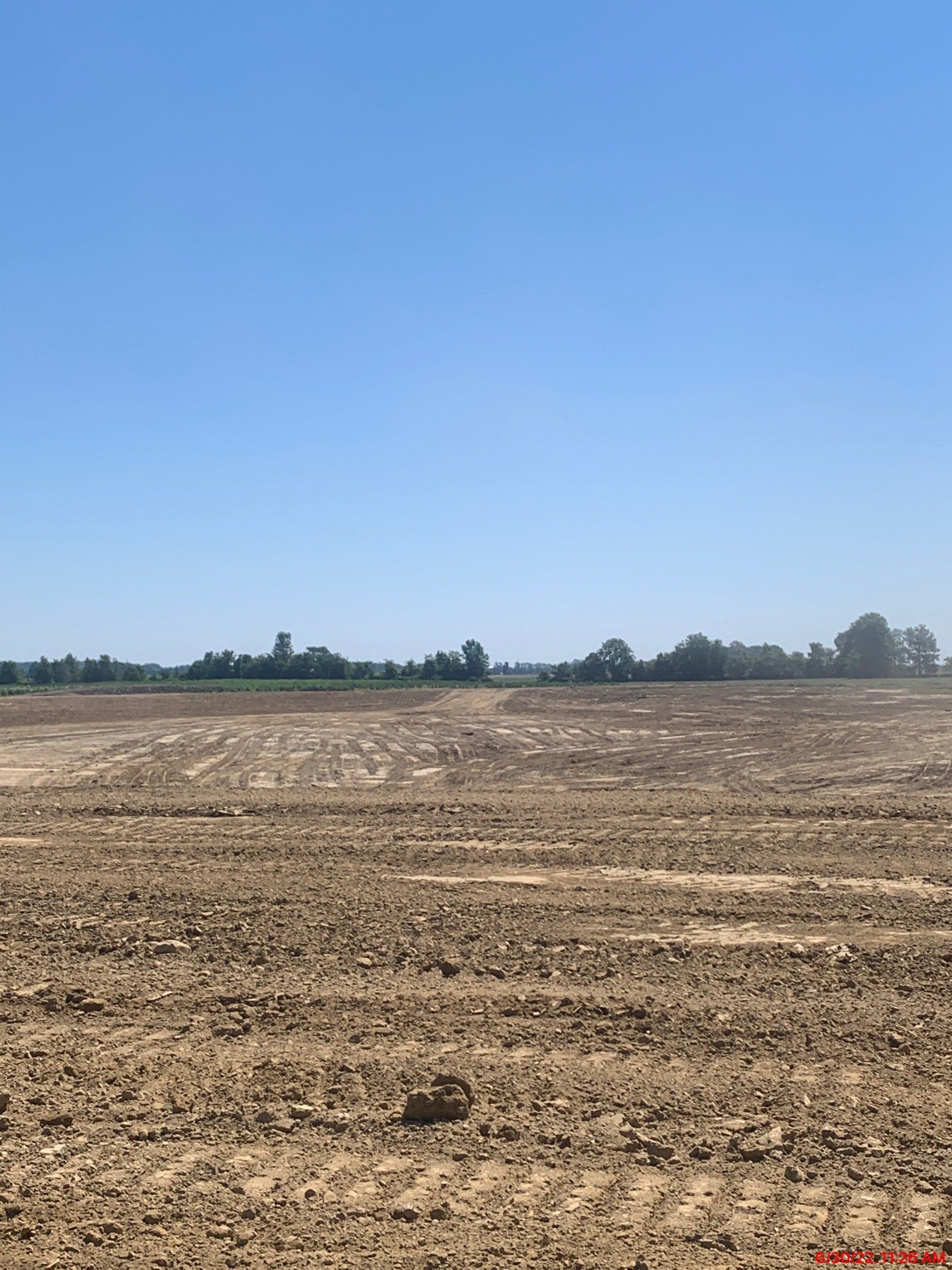 A barren, tilled field under a clear, blue sky. Distant trees and a structure visible on the horizon.