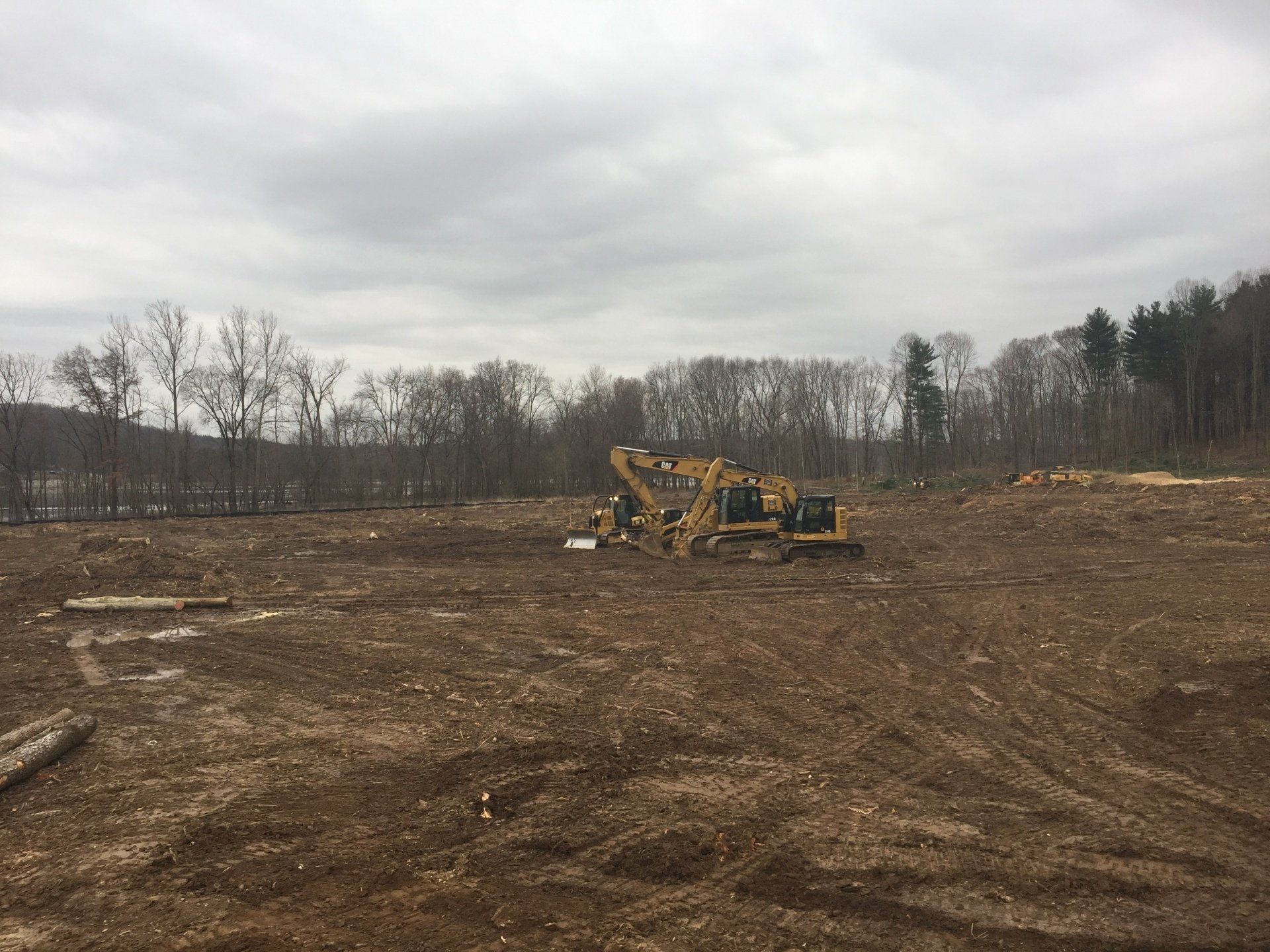 Earth-moving equipment on a cleared, muddy field, trees in the background under an overcast sky.