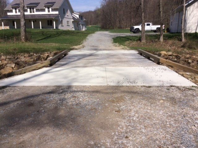 Concrete bridge over a gravel drive leading to a house, with a truck parked in the distance.