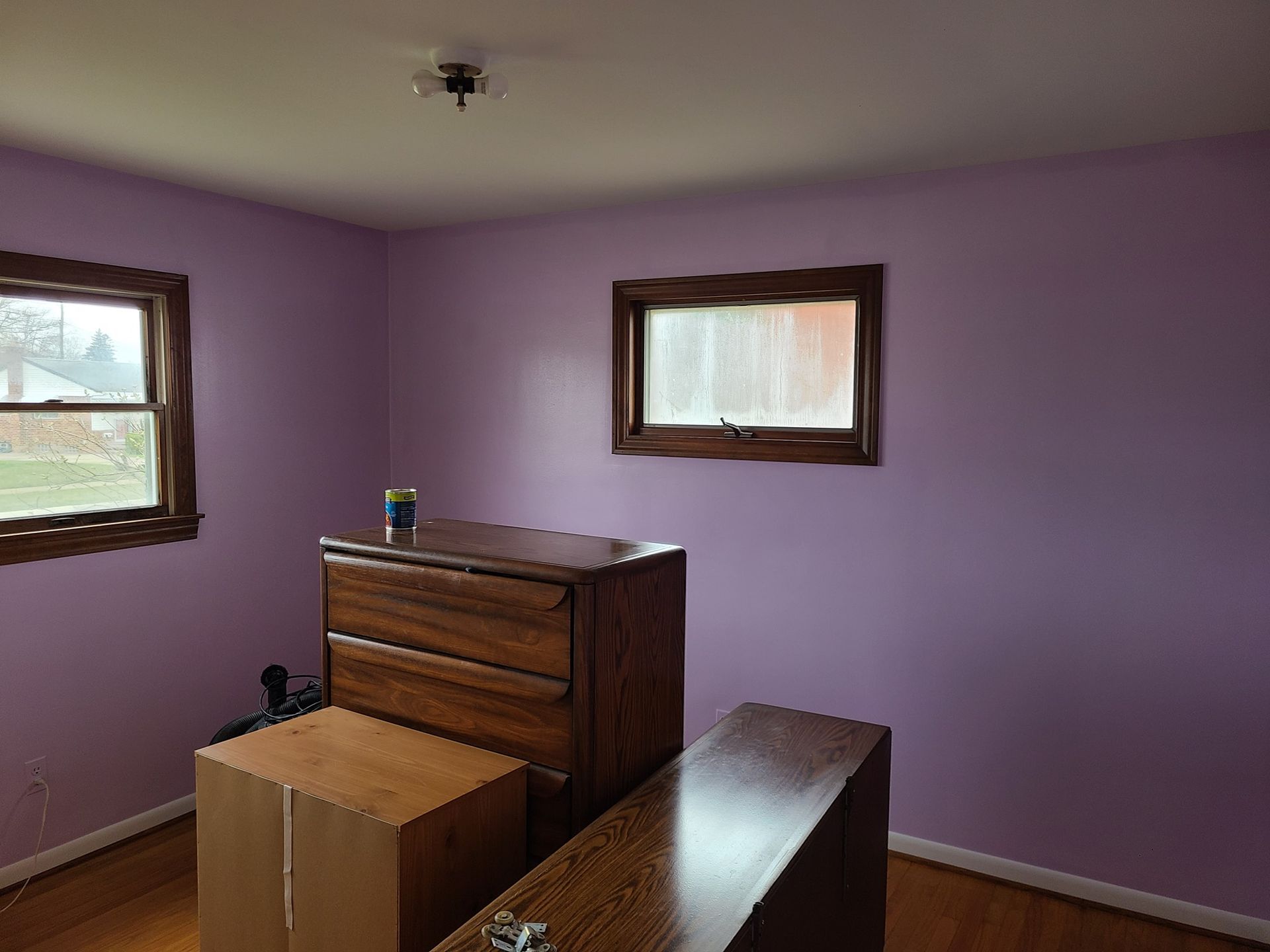 A room with purple walls , a dresser , and two windows.
