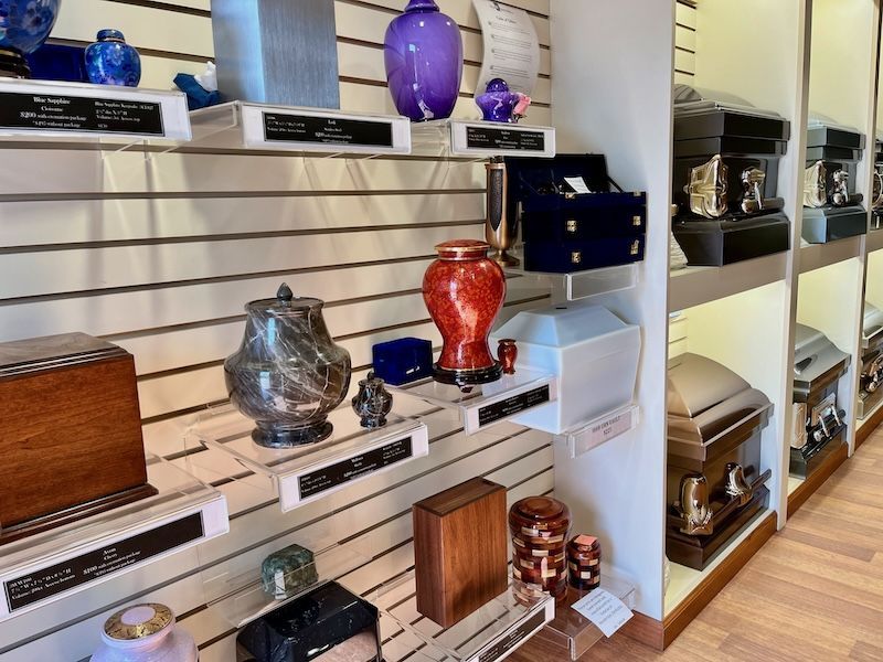 Shelves displaying cremation urns and boxes in a funeral home. Various shapes, sizes, colors and materials are shown.