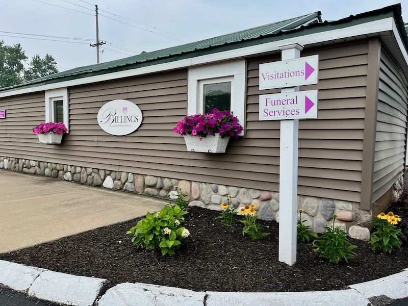 Exterior of a funeral home with signs for visitation and funeral services. Brown building with flowers.