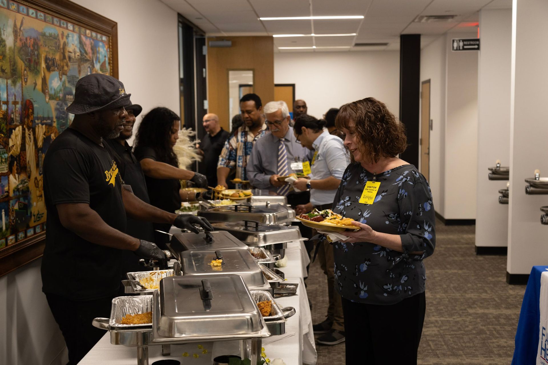 People at a buffet, served by staff. Food is in warming trays. The setting is indoors.