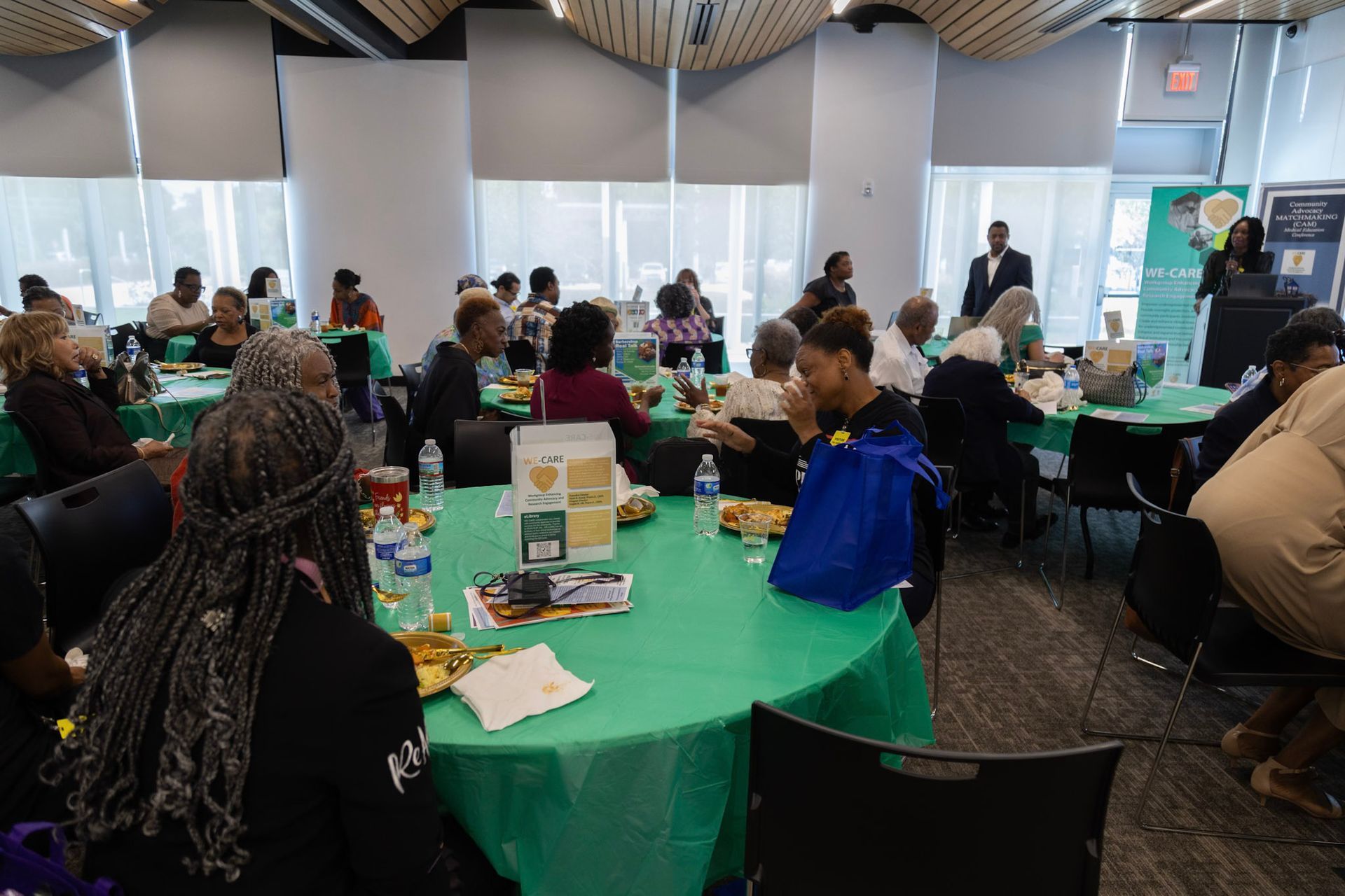 People seated at round tables with green tablecloths in a large room, listening to a presentation.
