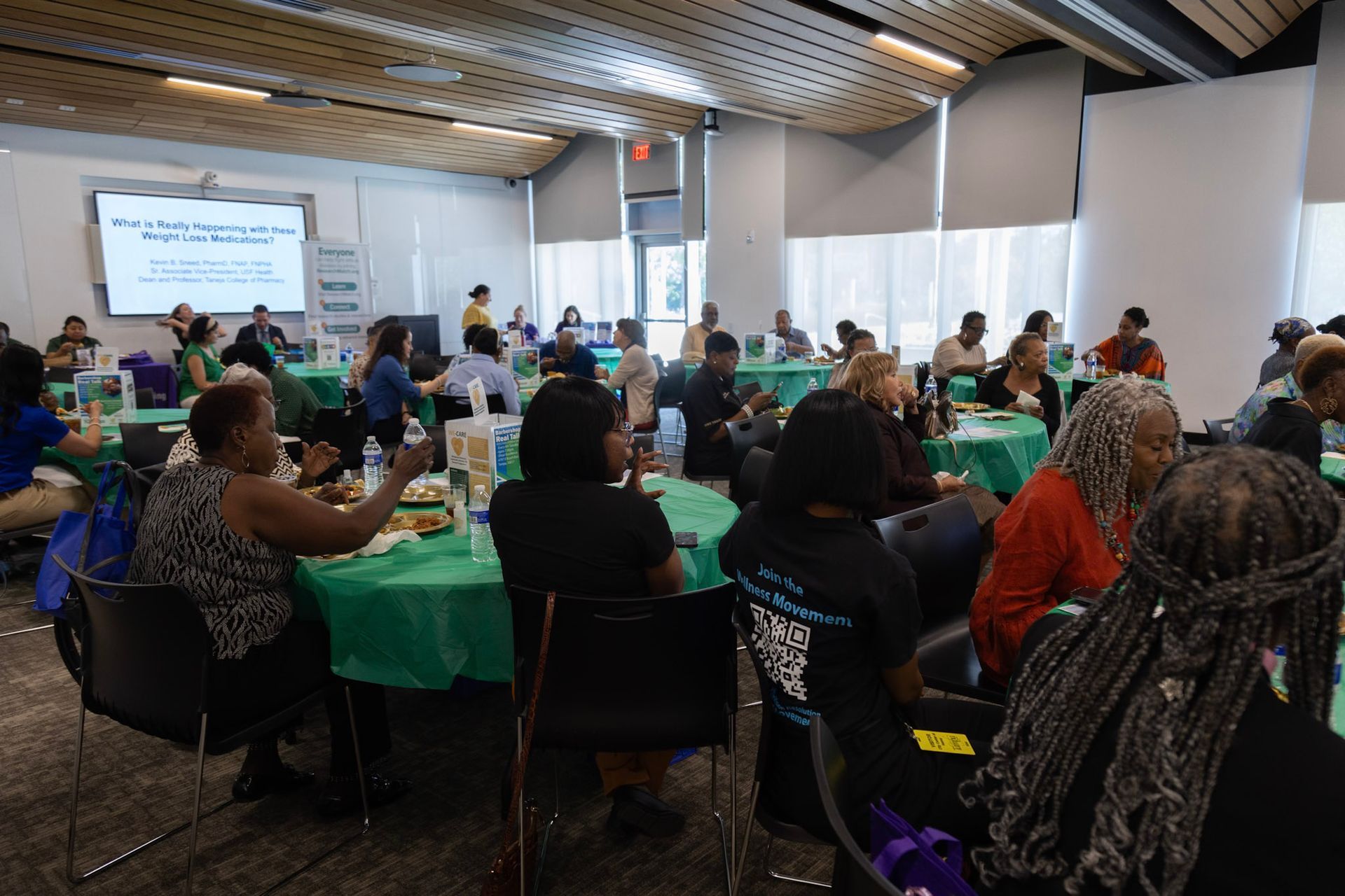 People seated at tables in a room with green tablecloths, attending an event with a screen and windows.