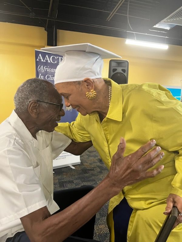 Woman in yellow embraces an older person, both smiling. The woman wears a mortarboard hat. Indoor setting.