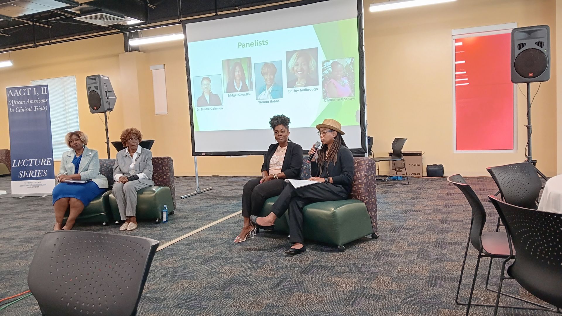 Panel discussion in a conference room with a screen displaying headshots. Speakers and audience seating are present.
