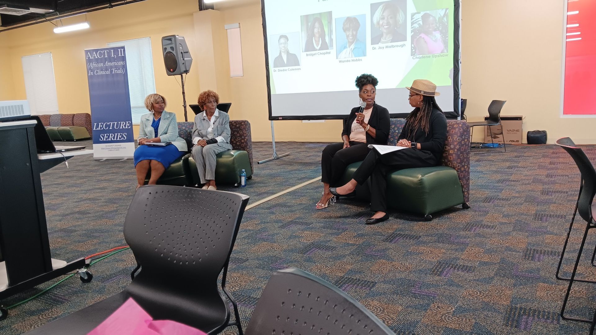 Panel of four people seated on chairs, speaking in a conference room with a screen displaying headshots.