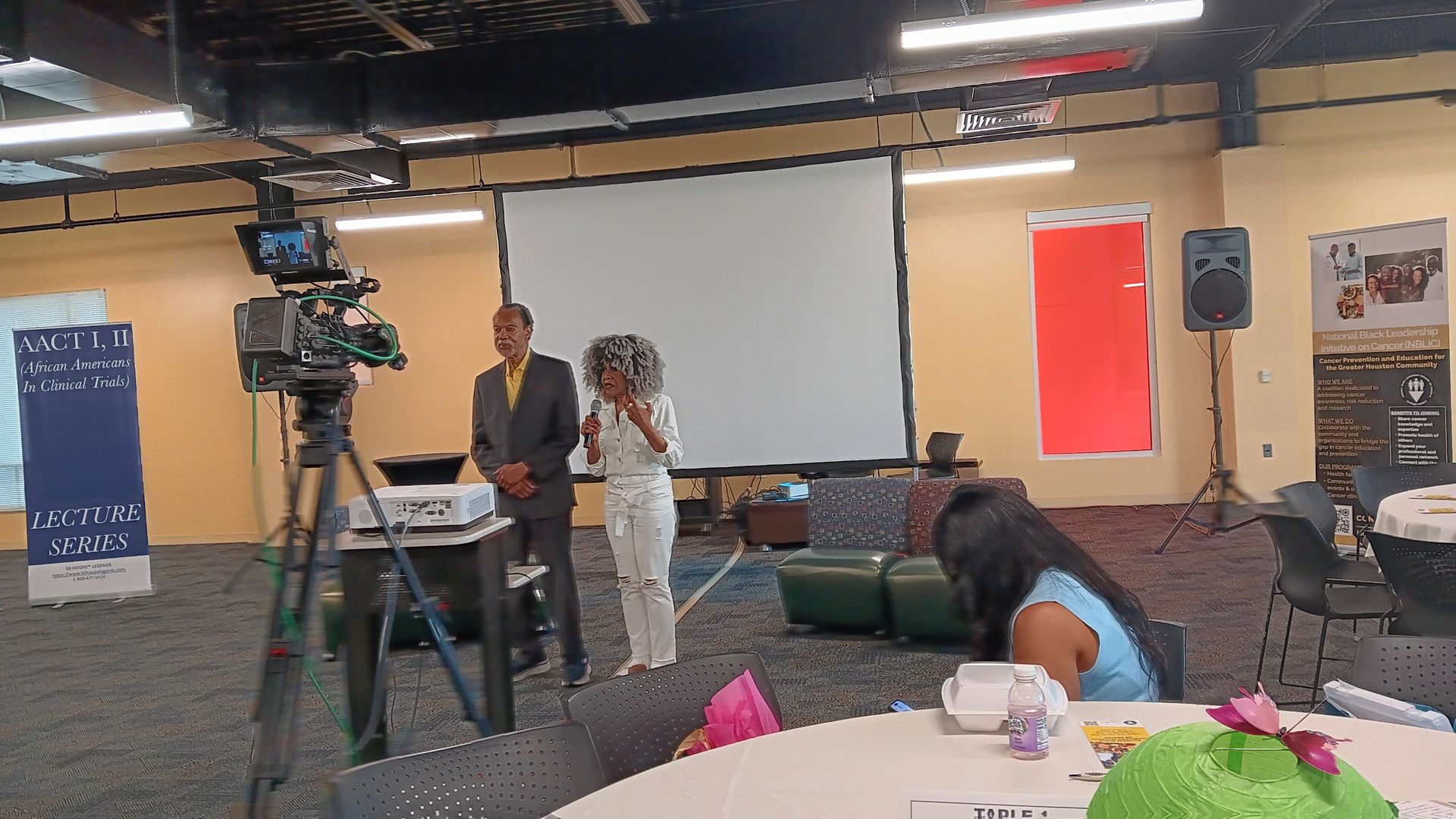 Two people stand in front of a screen, likely presenting. A video camera, speaker, and attendees are visible.