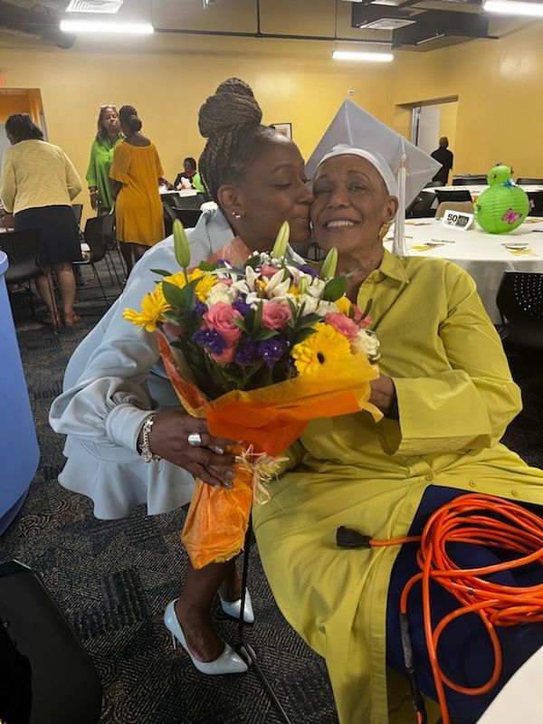 Woman kisses a graduate holding flowers, both smiling. Graduation ceremony setting. Yellow, white, and orange colors.