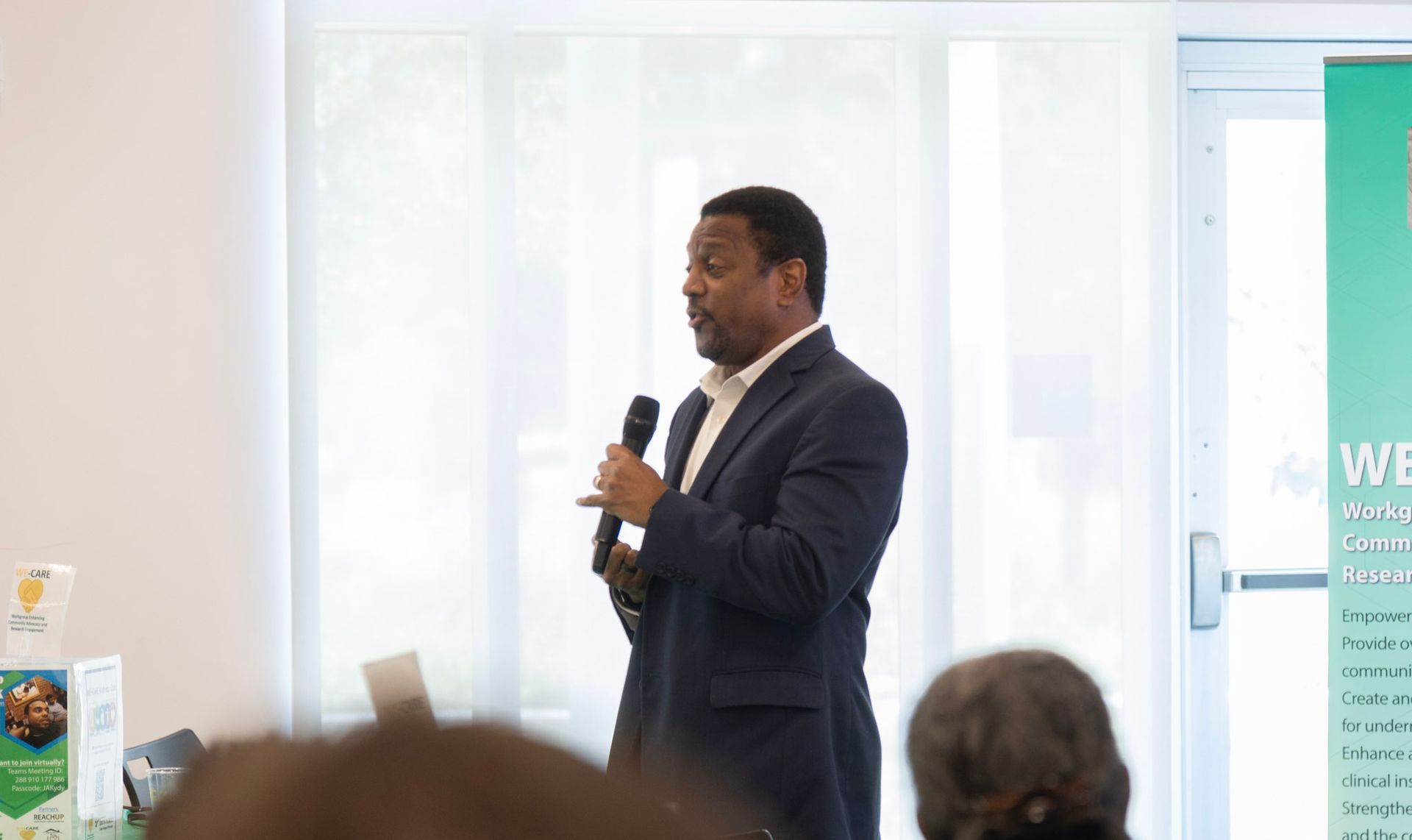 Man in a blue suit speaks into a microphone in a well-lit room with windows.