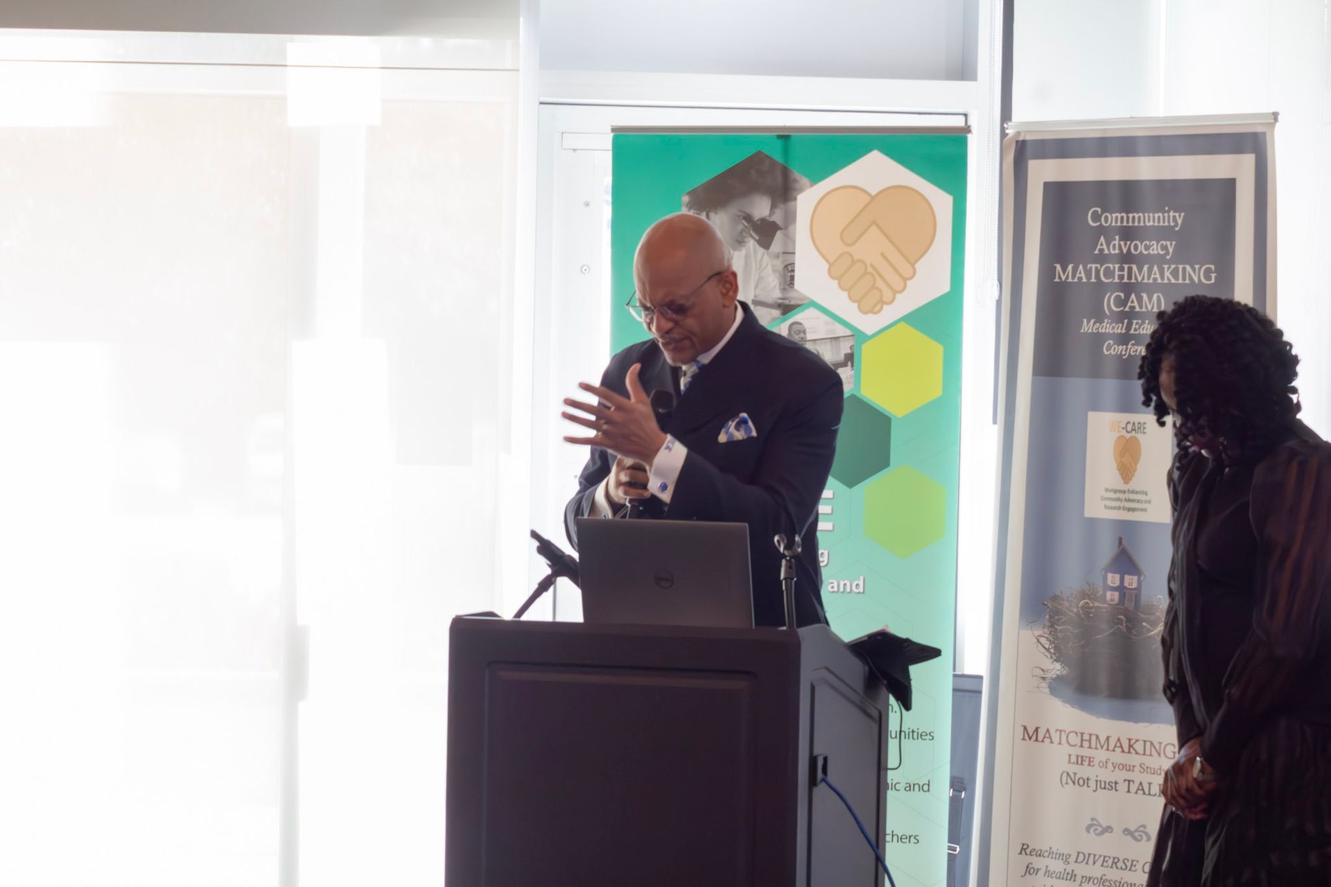 Man speaking at a podium with a laptop, gesturing. Woman stands to the side near a banner.
