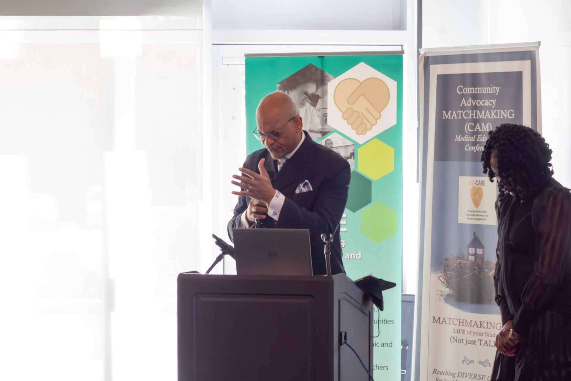 Man speaking at podium with laptop, green backdrop, woman in dark attire on right.