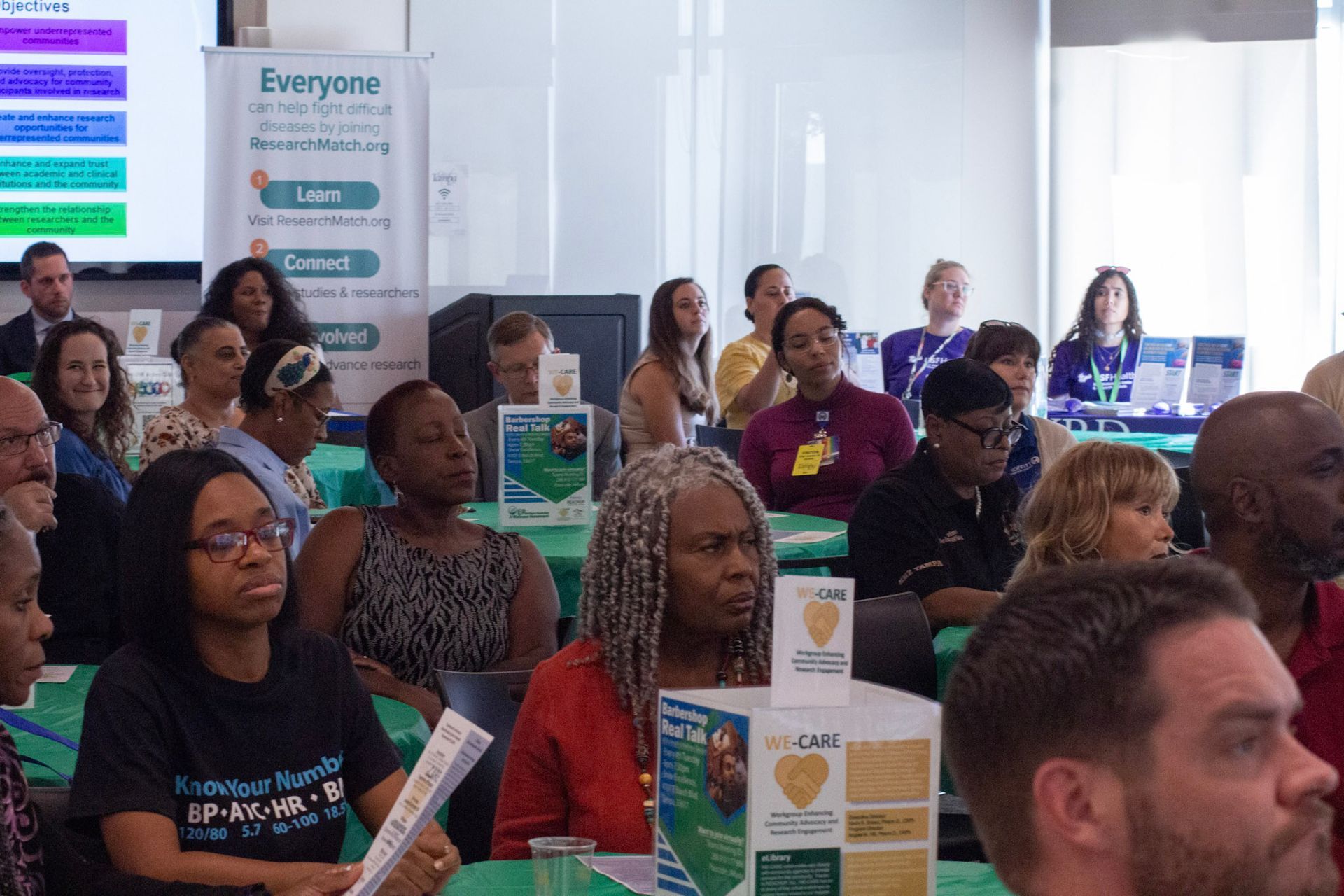 Audience in a room listening to a presentation; some looking toward the camera, some looking to the front.