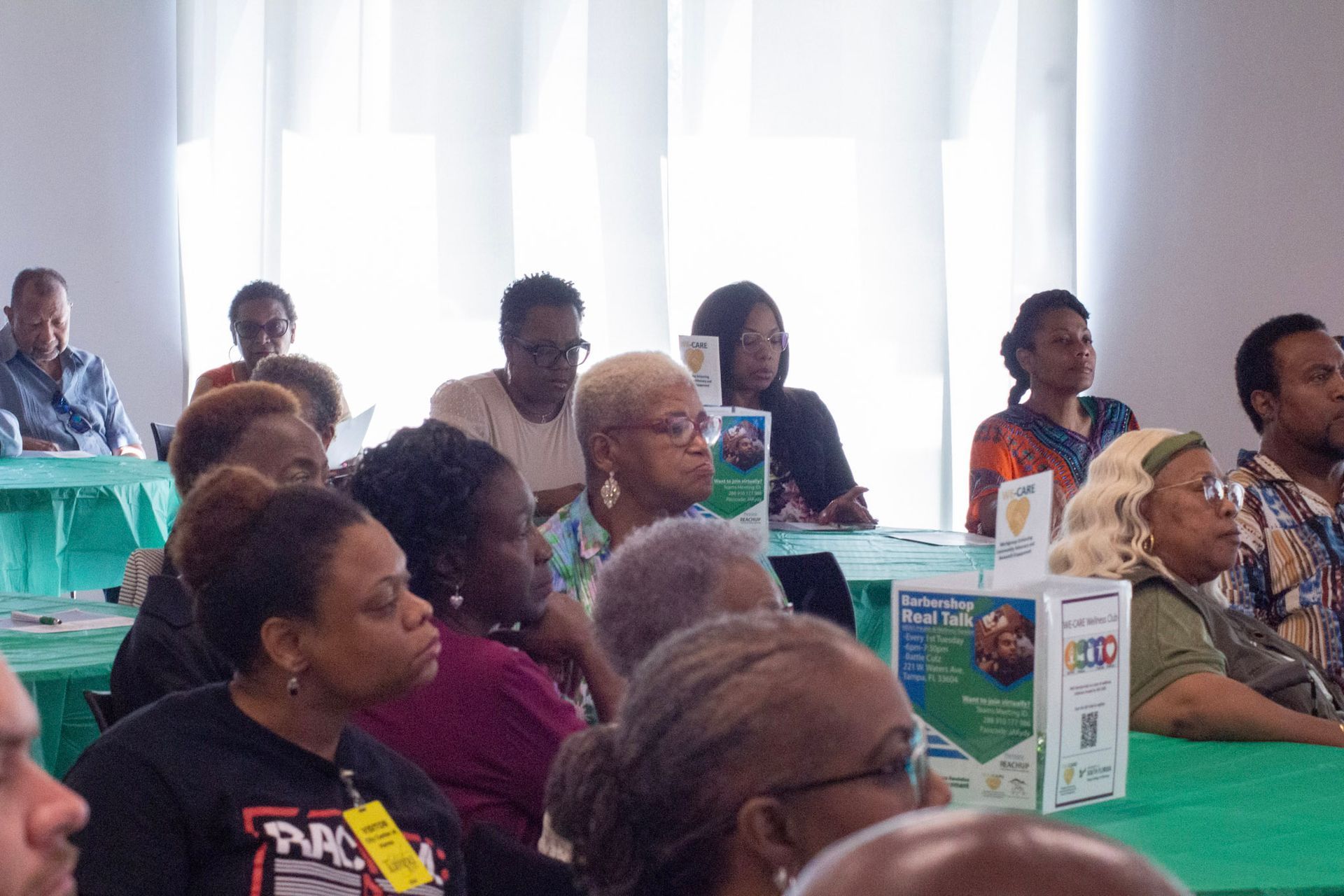 Audience at a conference, seated at tables. Several are looking forward, listening. Bright room with windows.