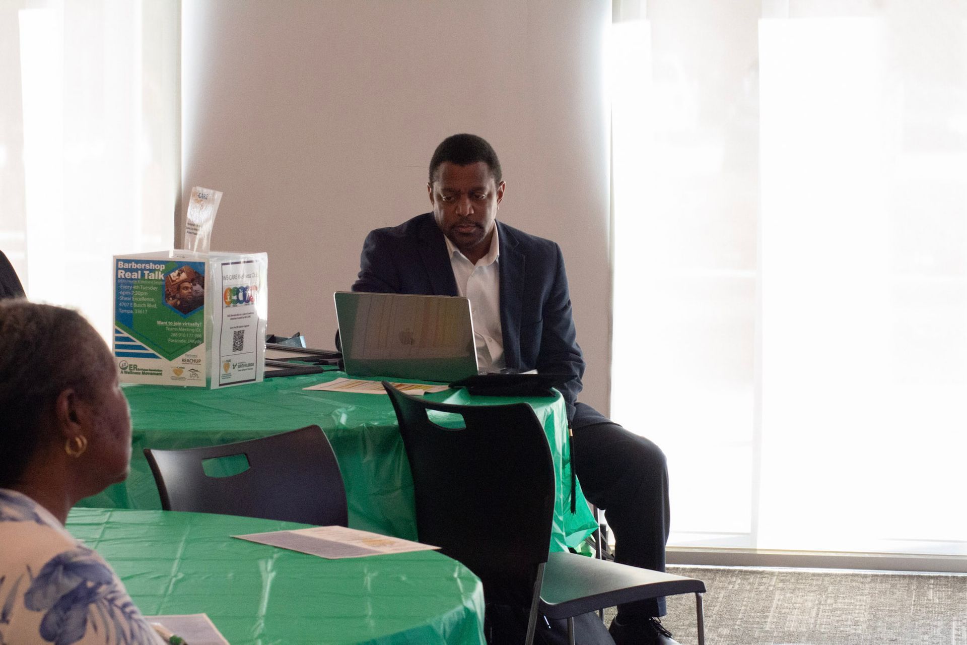 Man in suit using laptop at a table covered in green cloth; another person is seated nearby.