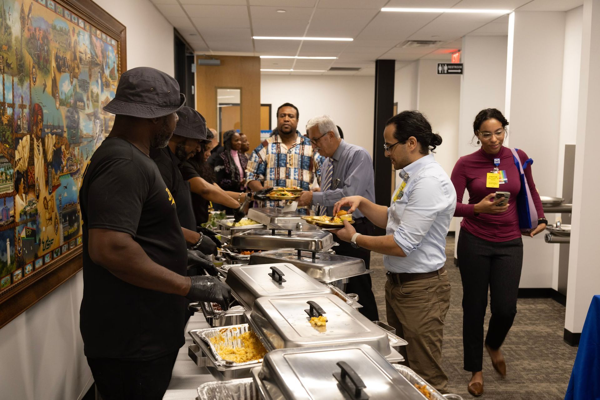 People serving themselves food from buffet line in a hallway; painting on the wall.