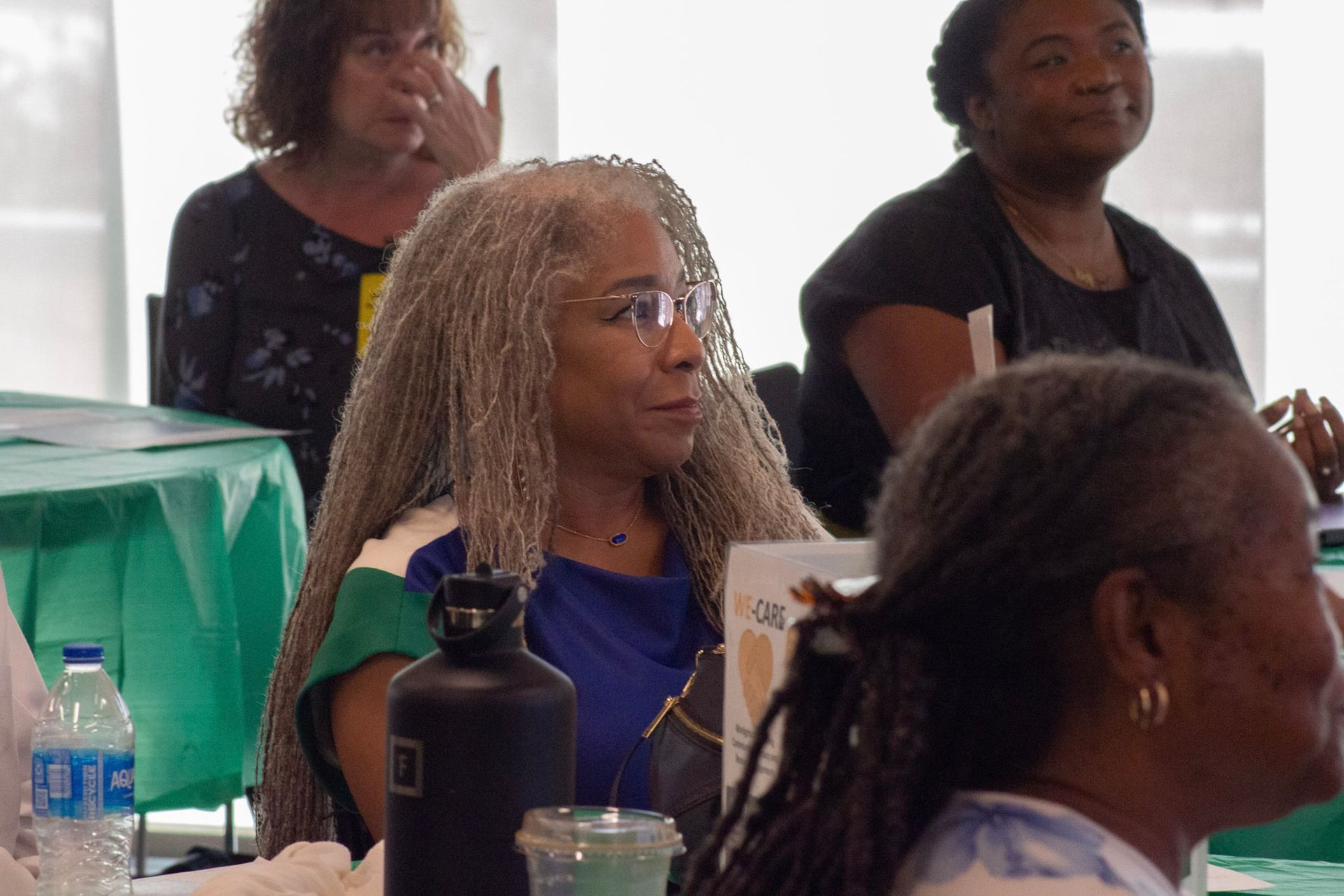 Woman with long gray hair and glasses at a table, looking to the side. Other people are in the background.
