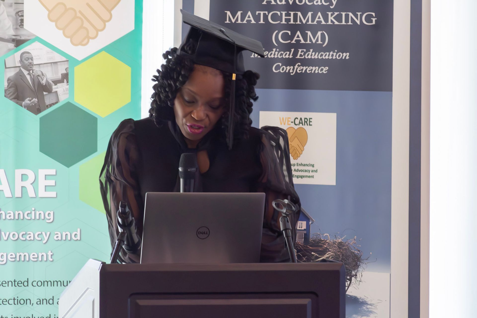 Woman speaking at podium with a laptop, wearing a graduation cap. Conference backdrop in background.