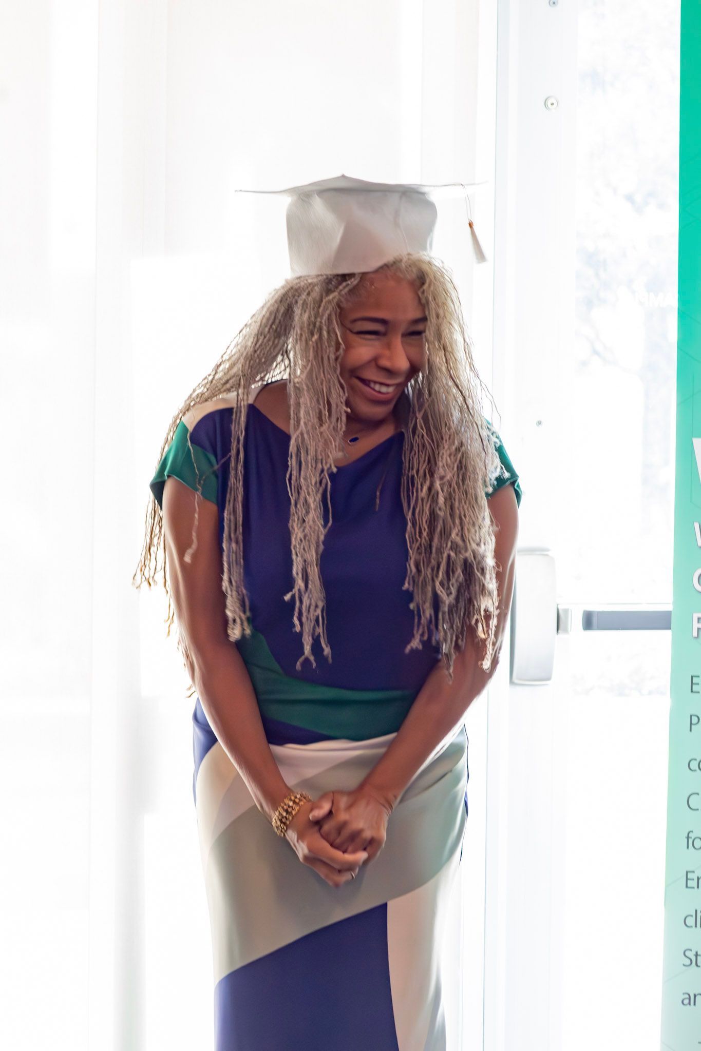 Woman wearing a graduation cap, smiling. She wears a blue, green, and cream dress, standing indoors by a window.
