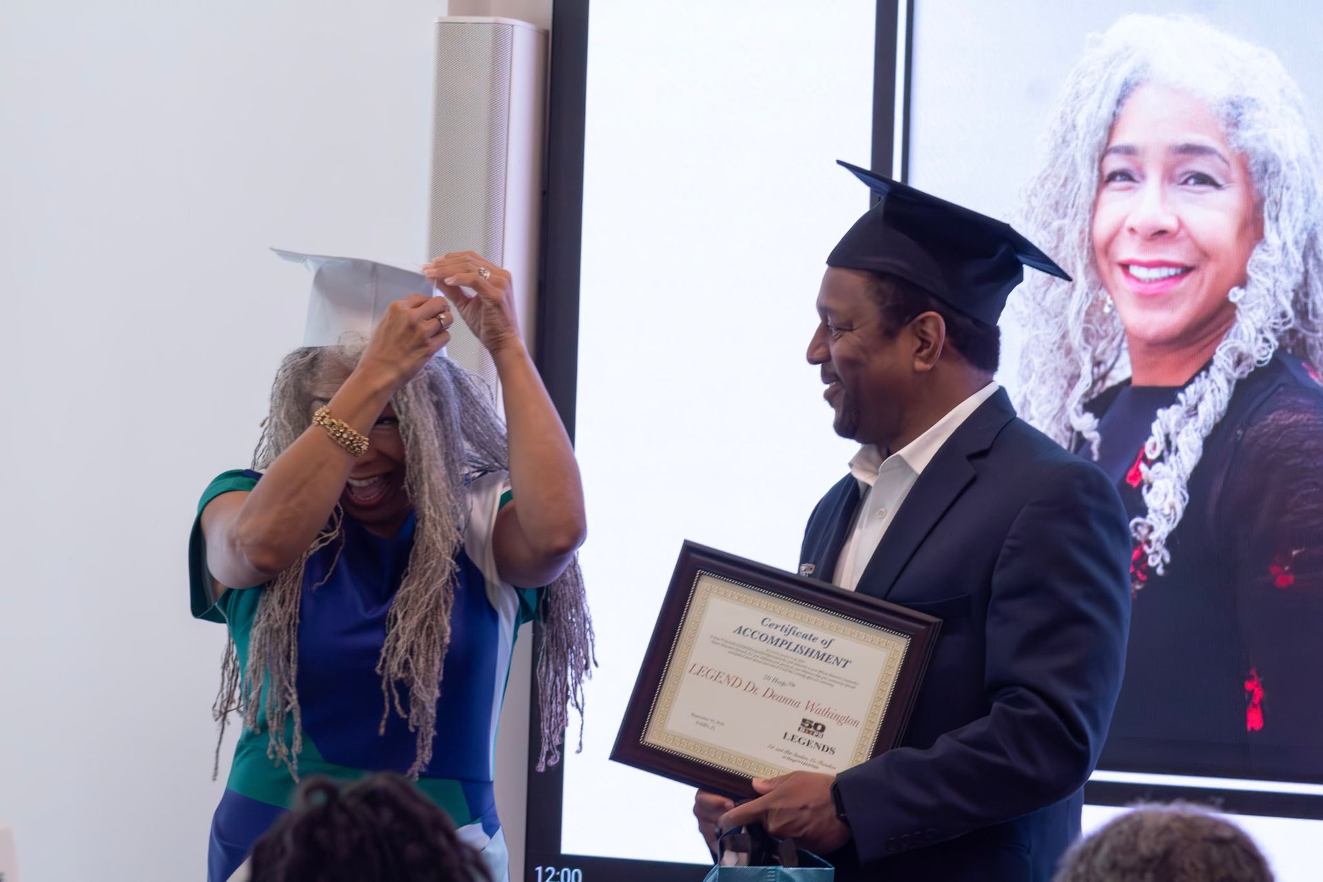Woman adjusting a graduation cap, man holding certificate, in front of a screen with a woman's portrait.