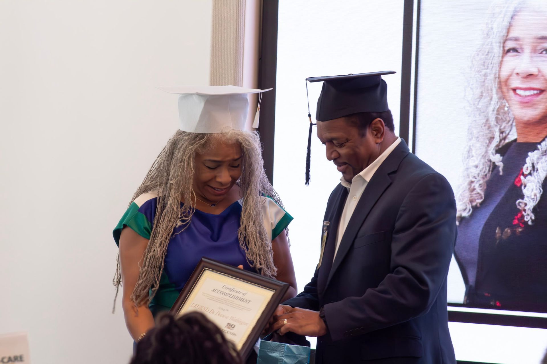 A person in a graduation cap accepts a certificate from another person in a suit, both smiling.