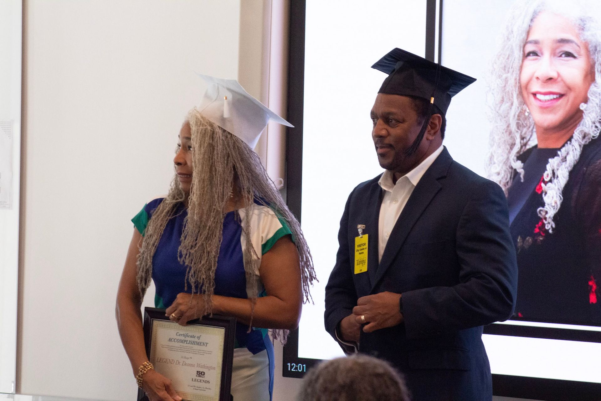 Two people in graduation caps stand before a screen; woman holds a certificate.