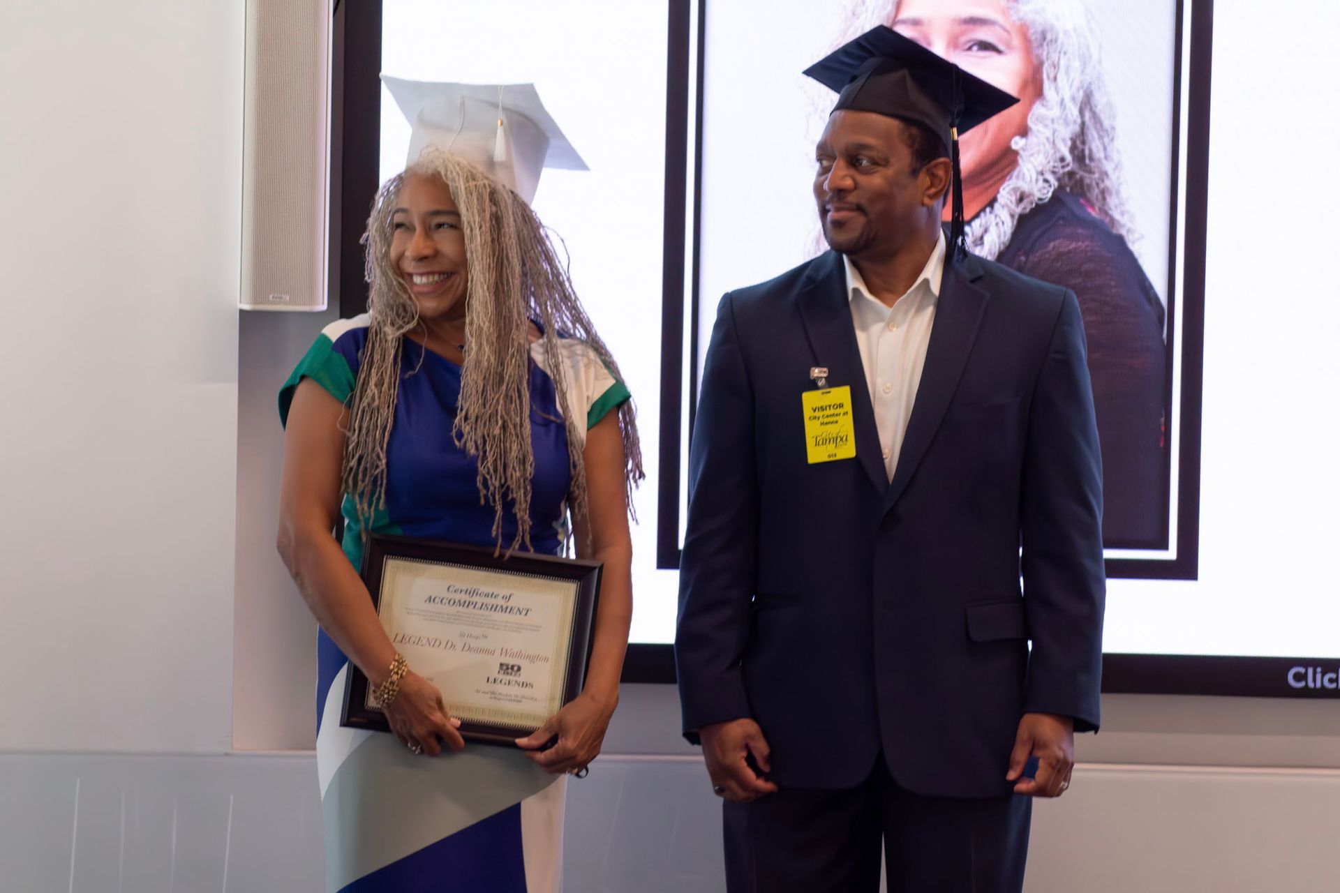 Two people in graduation caps, holding diplomas. Woman in blue dress smiles, man in suit looks to her.