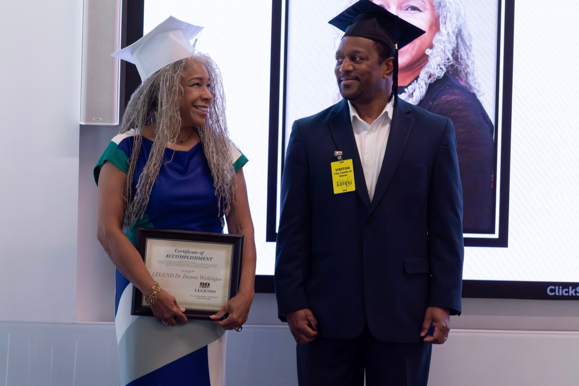 Woman in cap and gown holds a diploma, man beside her in a suit and cap smiles. Behind them a screen shows a headshot.