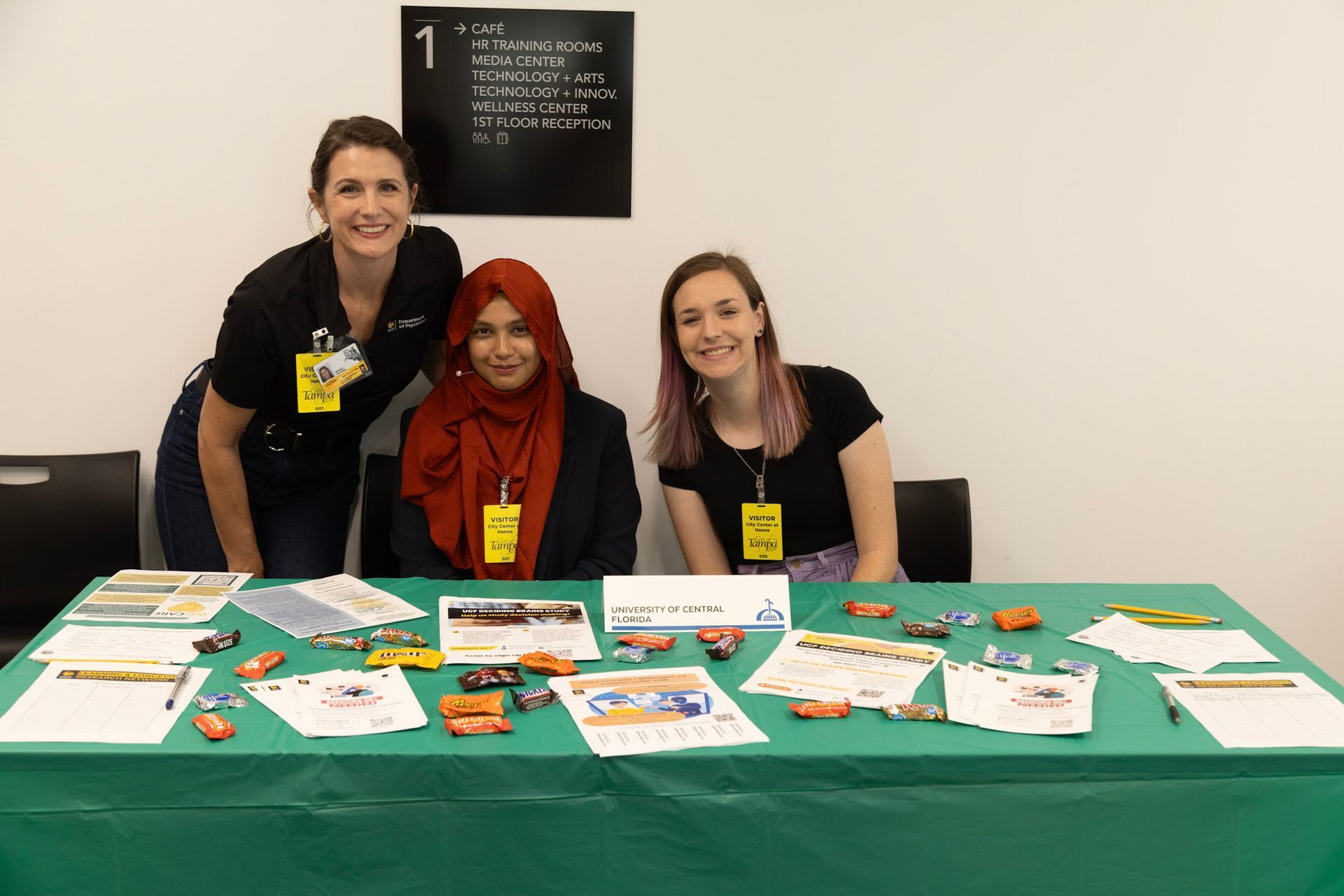 Three people at a table with brochures and model cars. Women smiling. Green tablecloth, white wall backdrop.