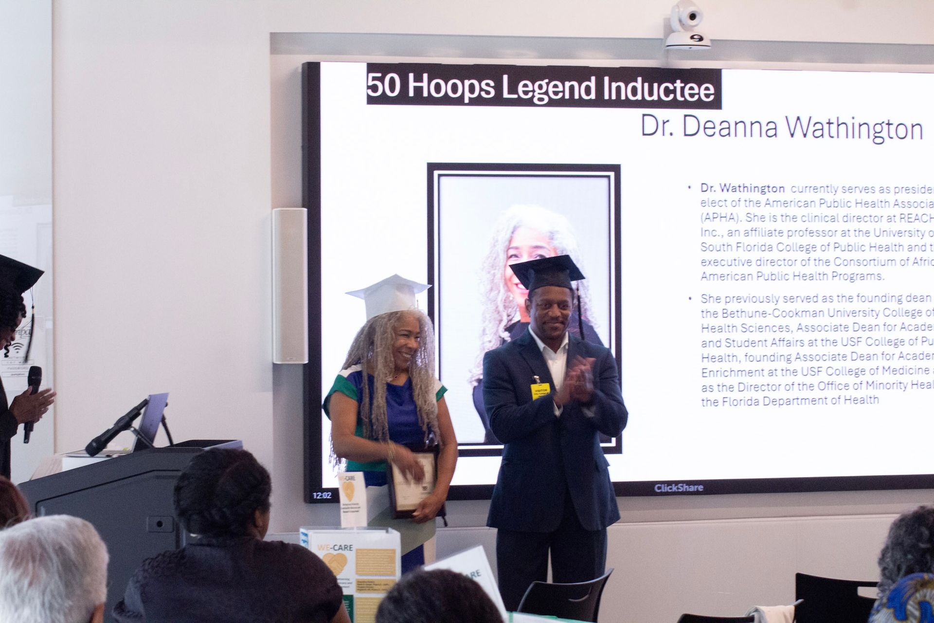 Two people clapping at a ceremony with a screen in the background.