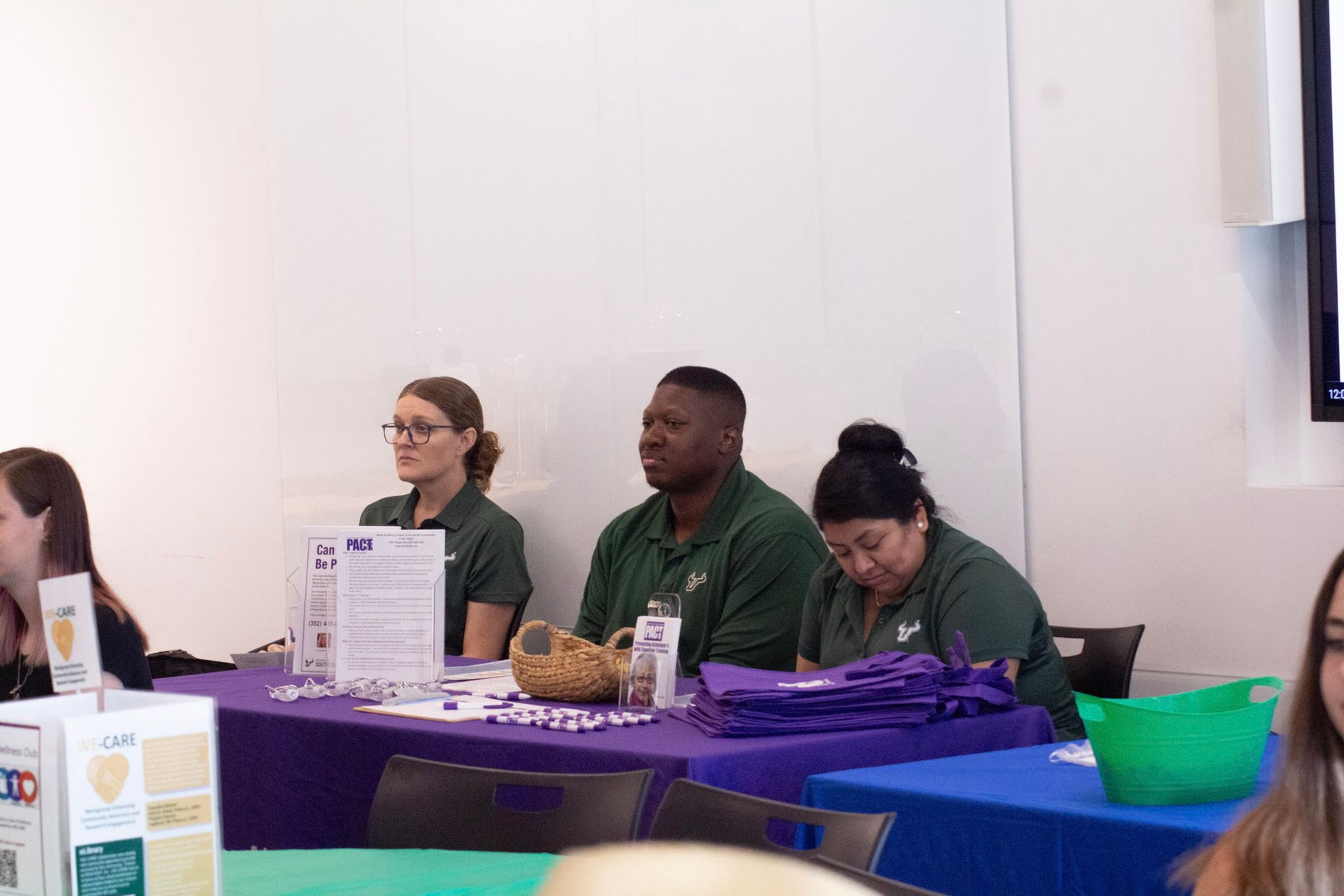 Three people at a table, wearing green shirts. Purple tablecloth, promotional materials, and a white wall in the background.
