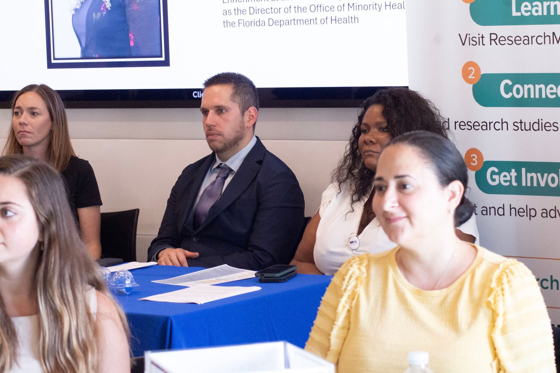 People seated at a table, likely in a meeting or presentation, with a blue tablecloth.