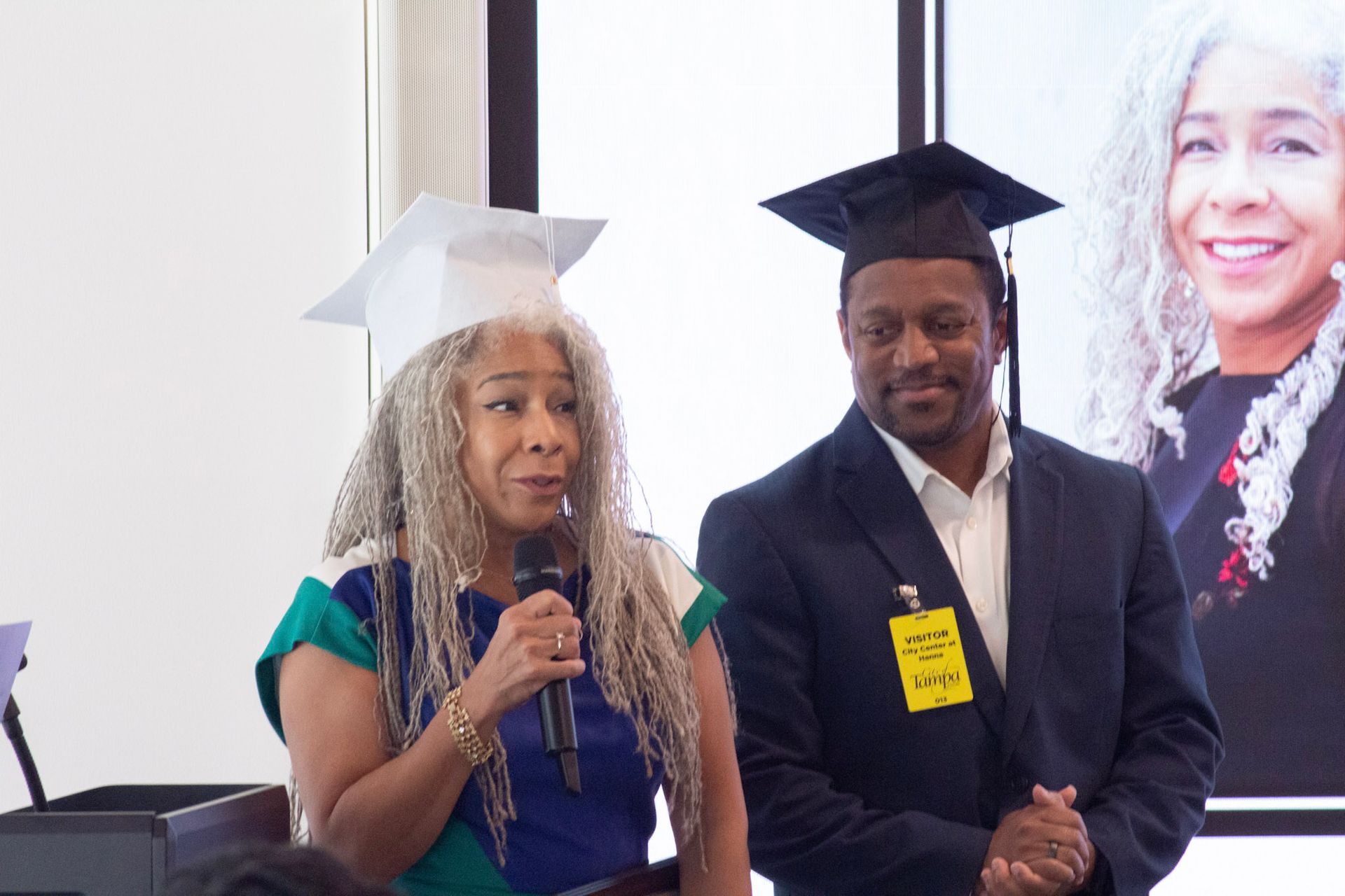 Woman in blue dress speaks into mic; man in suit smiles. Both wear graduation caps.