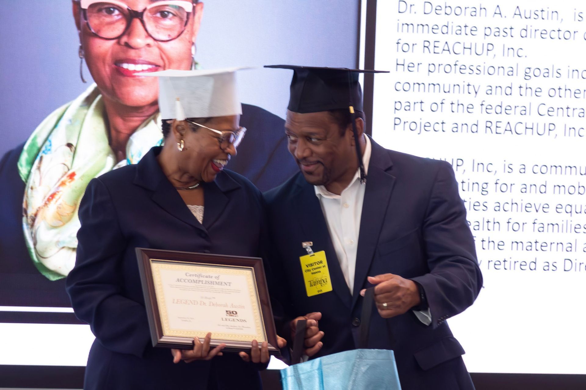 Two people, one in a graduation cap, holding a framed certificate and smiling. A photo of another woman is in the background.