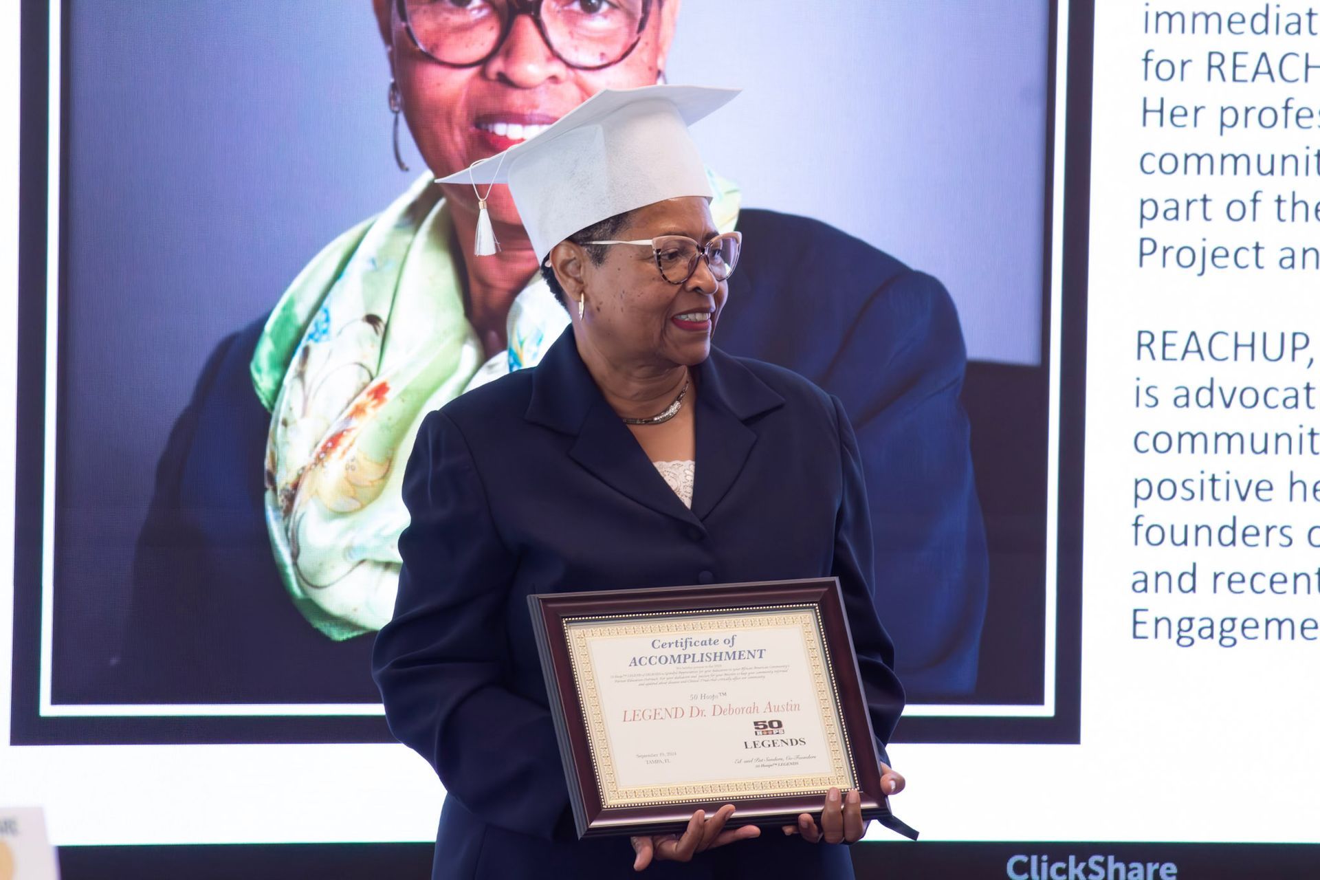 Woman in a graduation cap holding a certificate. A screen with text is behind her.