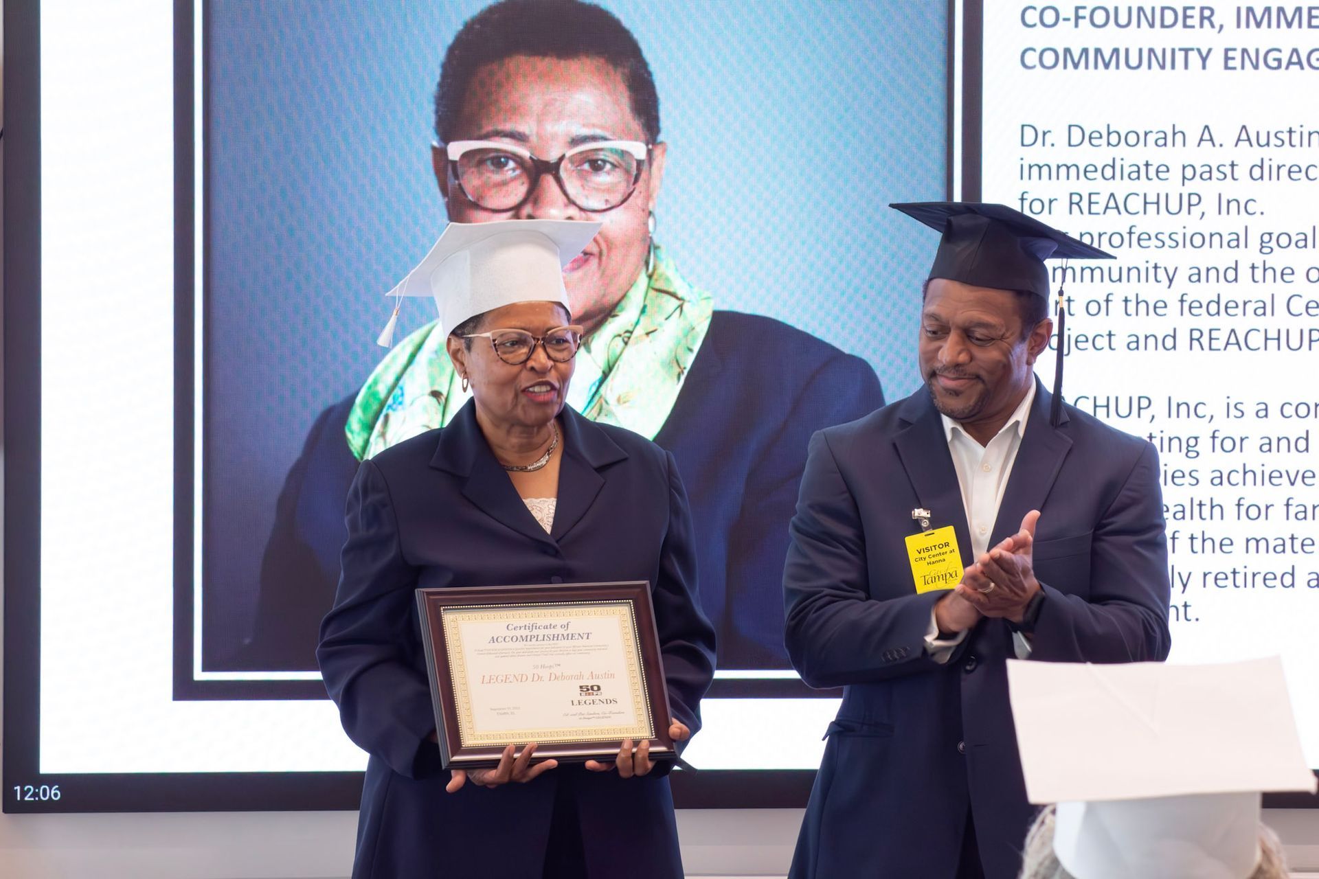 Two people, one holding a certificate, the other clapping, in front of a screen with a person's photo and text.