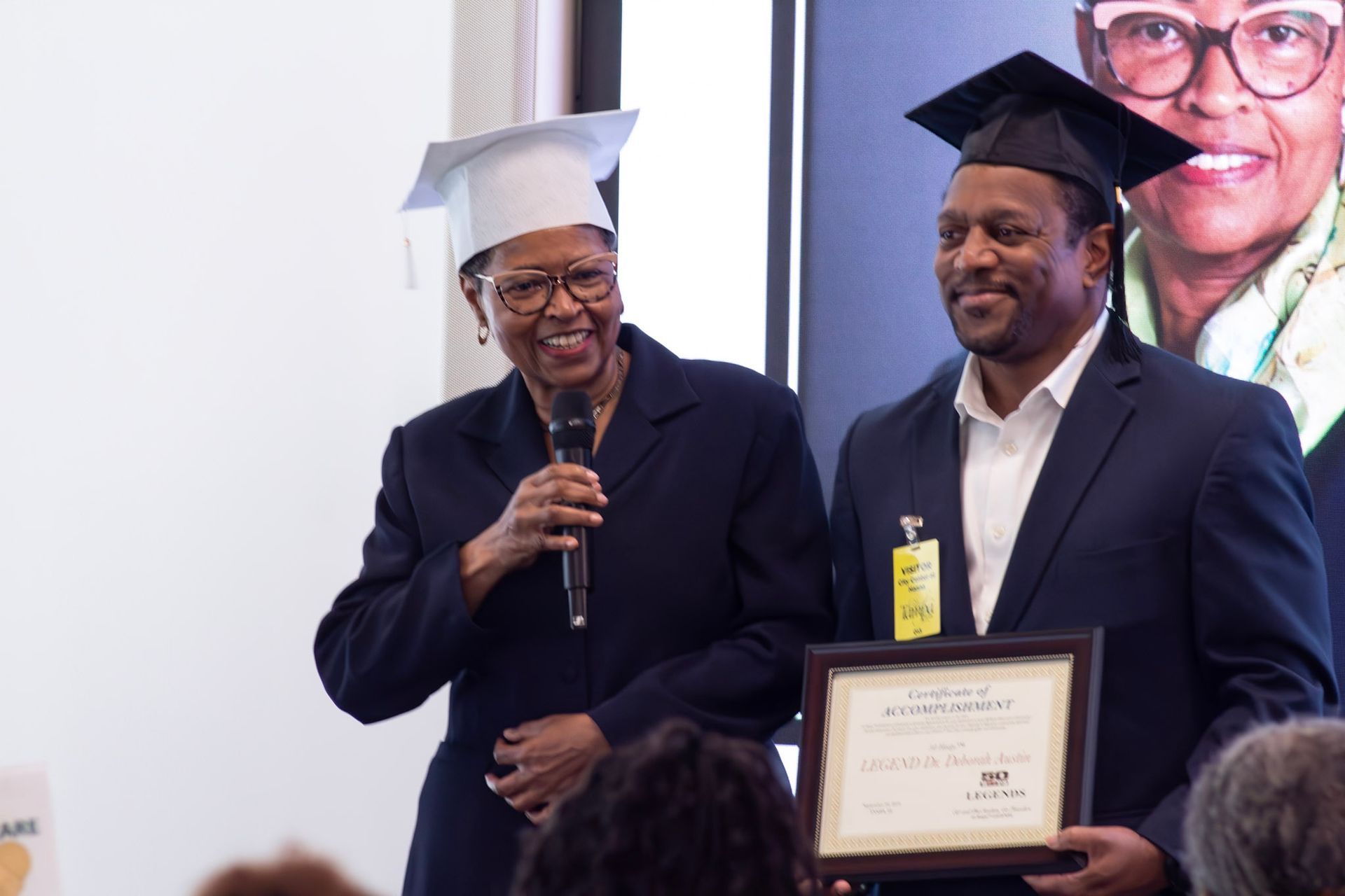 Woman with mic, holding diploma with man in graduation cap.
